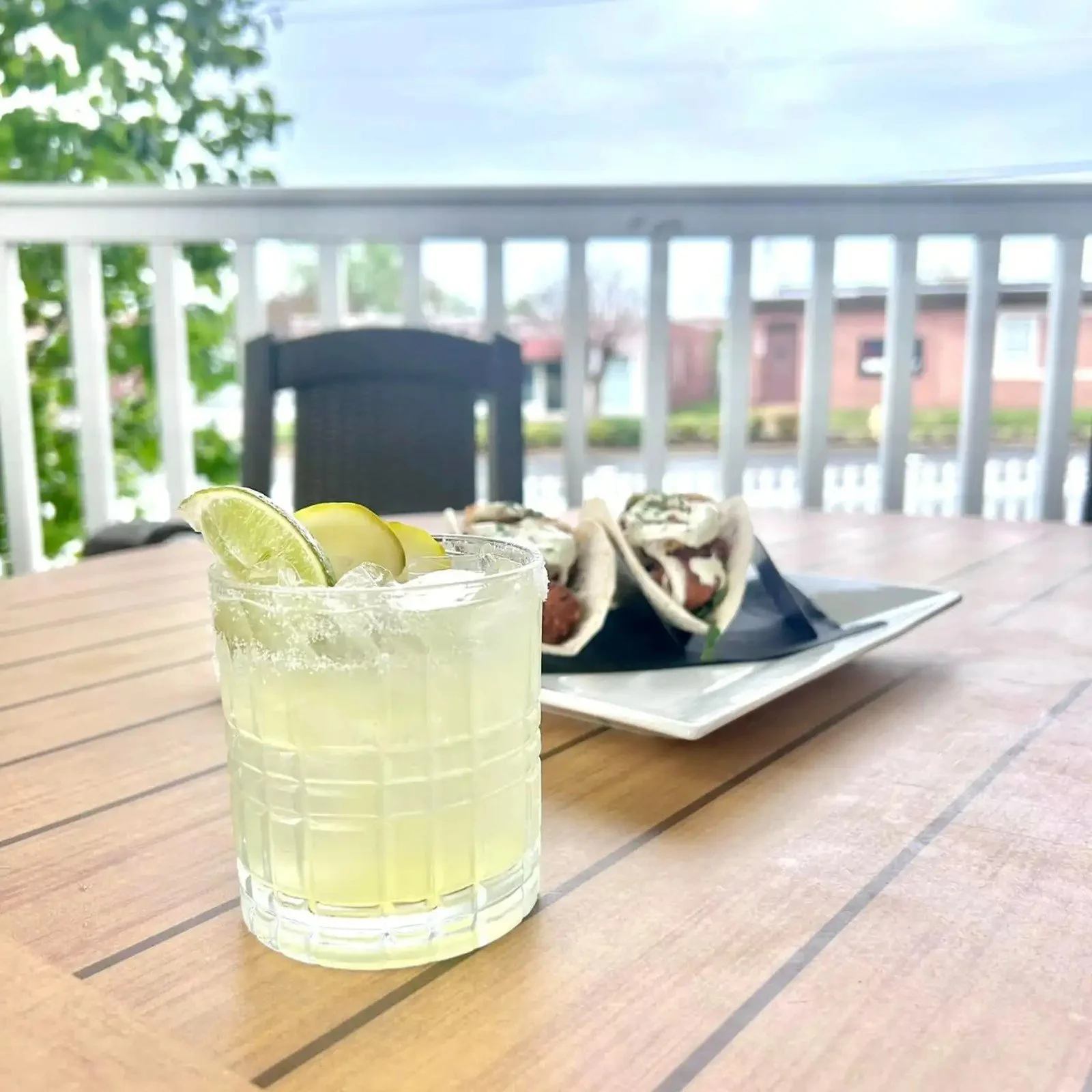 A glass of lemonade with slices of lime on a wooden table, with a plate of tacos in the background and a porch with a white railing and green trees outside.
