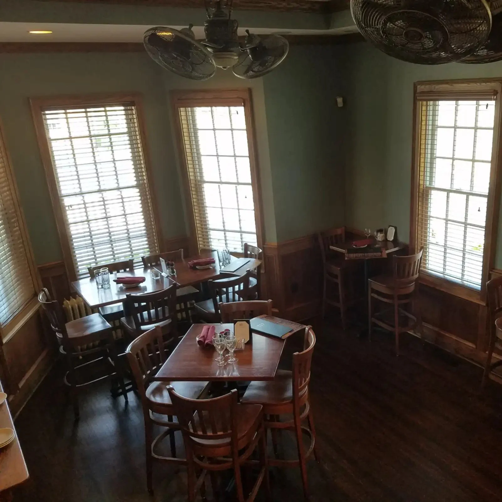 Interior of a restaurant dining area with several wooden tables and chairs, set with glasses, napkins, and menus. Large windows with wooden blinds, a ceiling fan, and dark wooden flooring.
