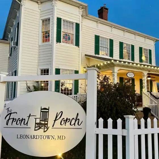 A large white two-story house with green shutters, decorated with string lights along the porch railing, and a white picket fence in front. A sign reads "The Front Porch, Leonardtown, MD."