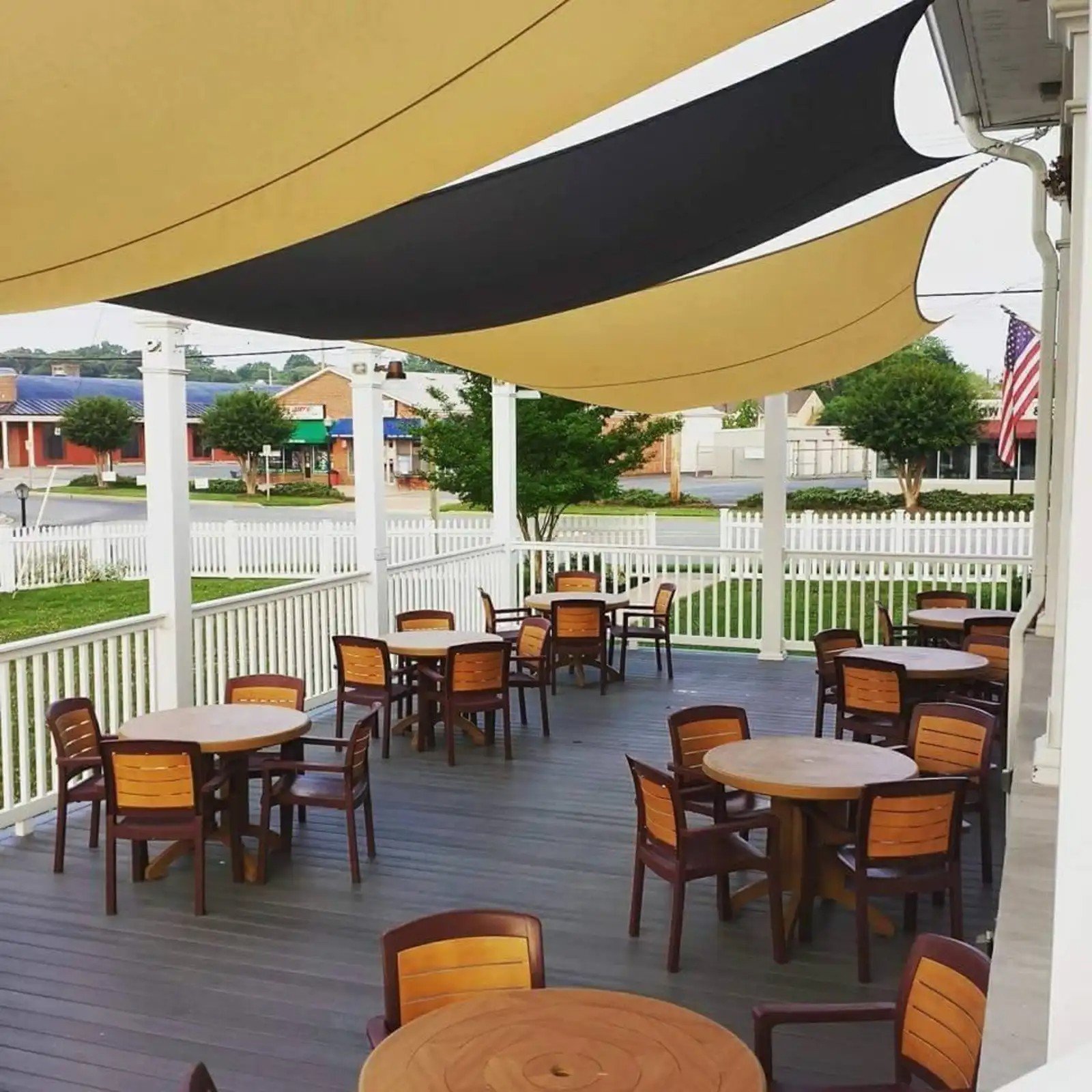 An outdoor patio area with wooden tables and chairs, shaded by large yellow and black fabric canopies, surrounded by a white picket fence, with trees, a street, and buildings in the background.