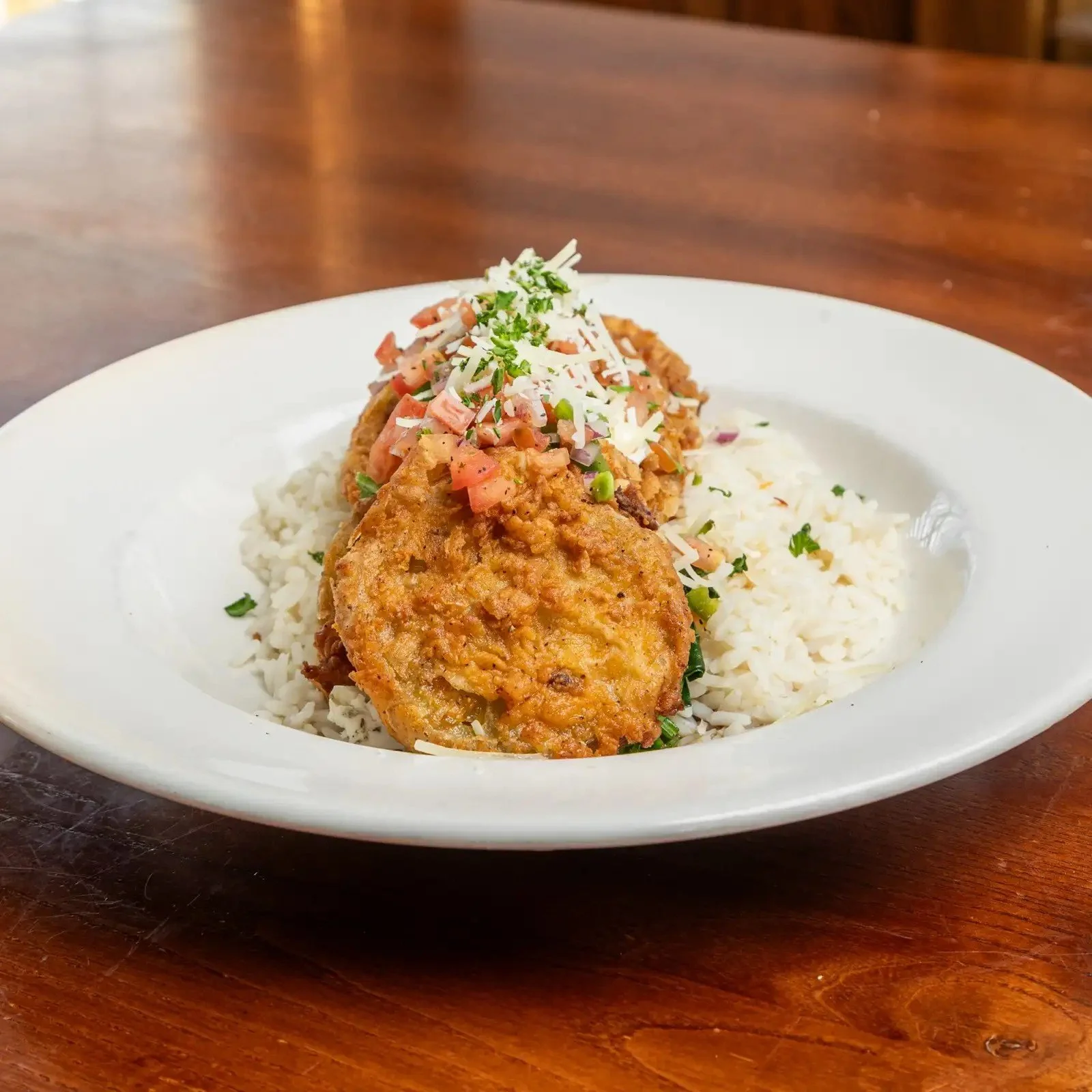 Plate of fried fish fillet served over white rice with diced tomatoes, onions, and herbs, garnished with grated cheese and chopped parsley.