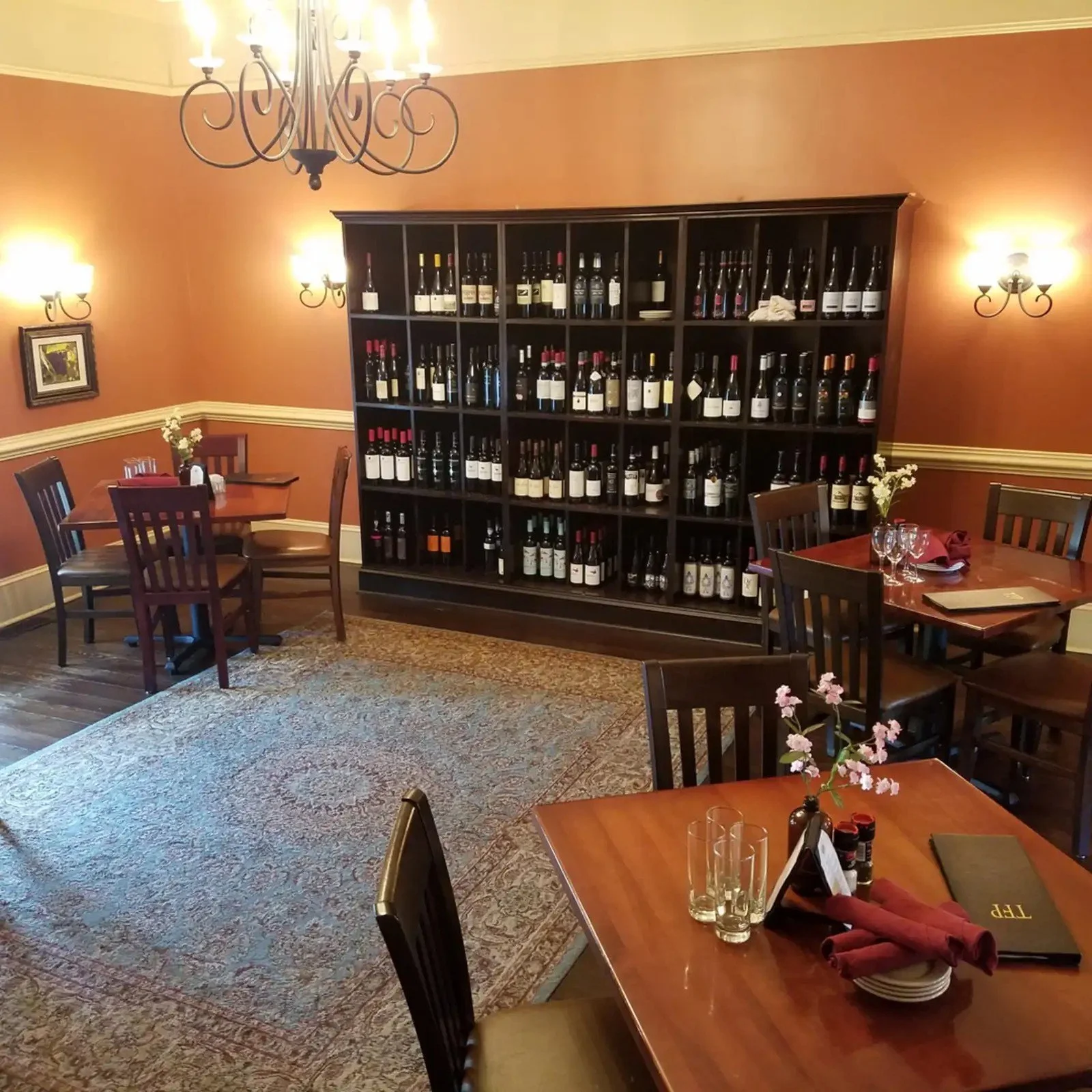 A cozy restaurant with a large wine rack against a salmon-colored wall, several tables with wine glasses, napkins, flowers, and menus, chandelier and wall sconces providing warm lighting, patterned carpet, and wooden chairs.