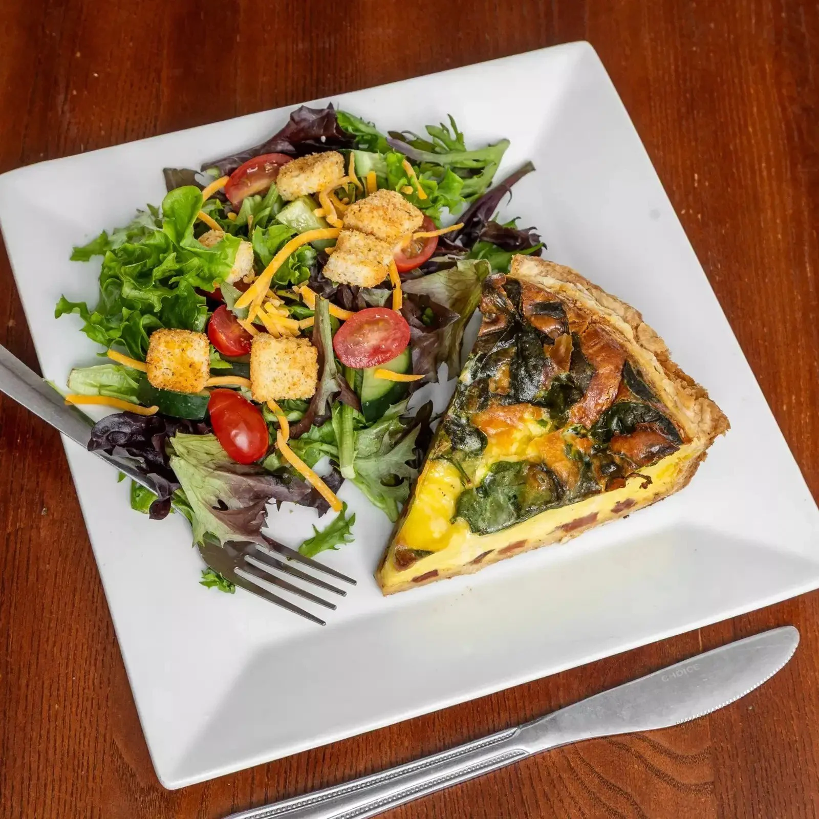 A white square plate with a salad on the left side and a slice of vegetable quiche on the right side, placed on a wooden table. The salad includes leafy greens, cherry tomatoes, cucumber, shredded cheese, croutons, and dressing.