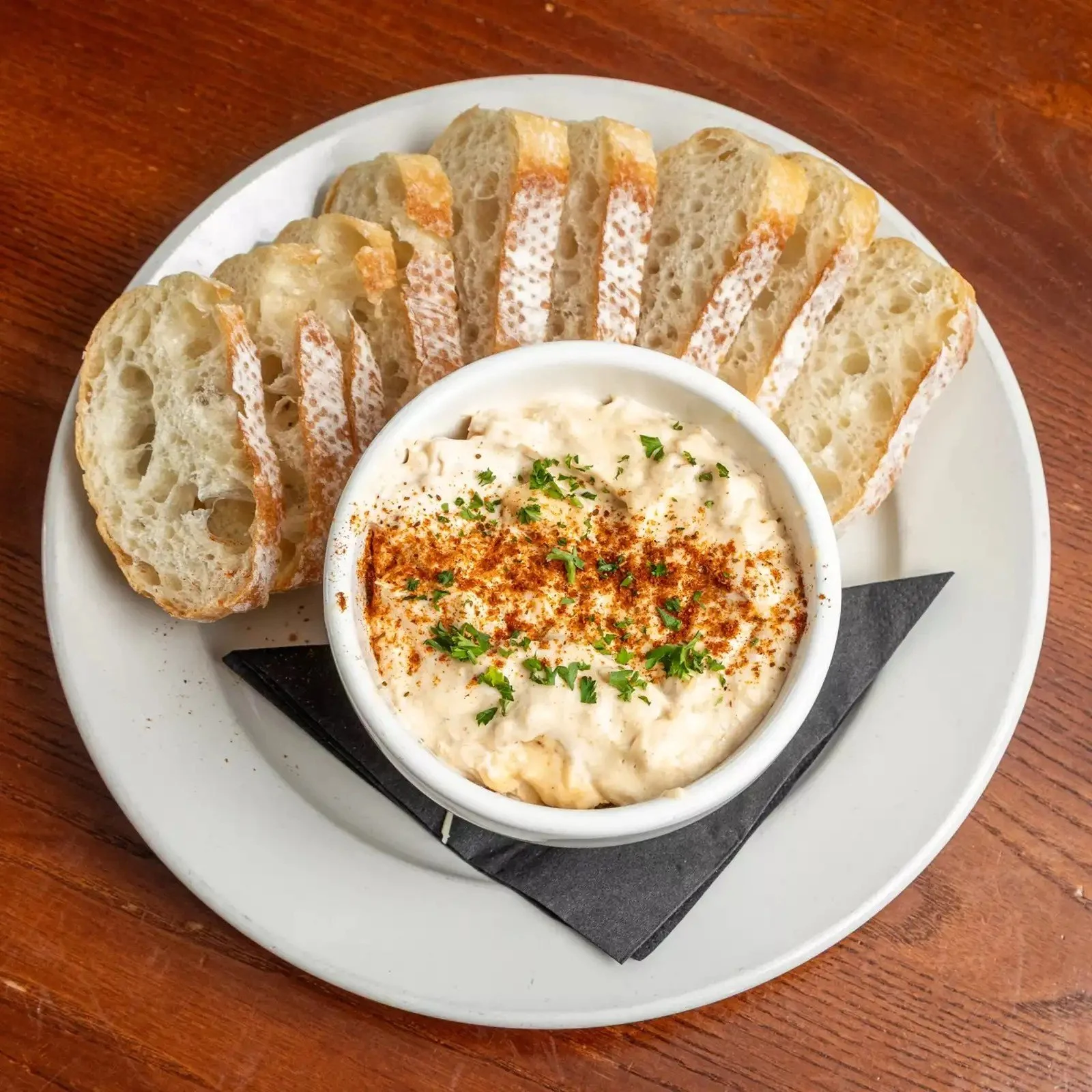 A white plate with sliced bread, a bowl of creamy dip topped with paprika and herbs, served on a wooden table.