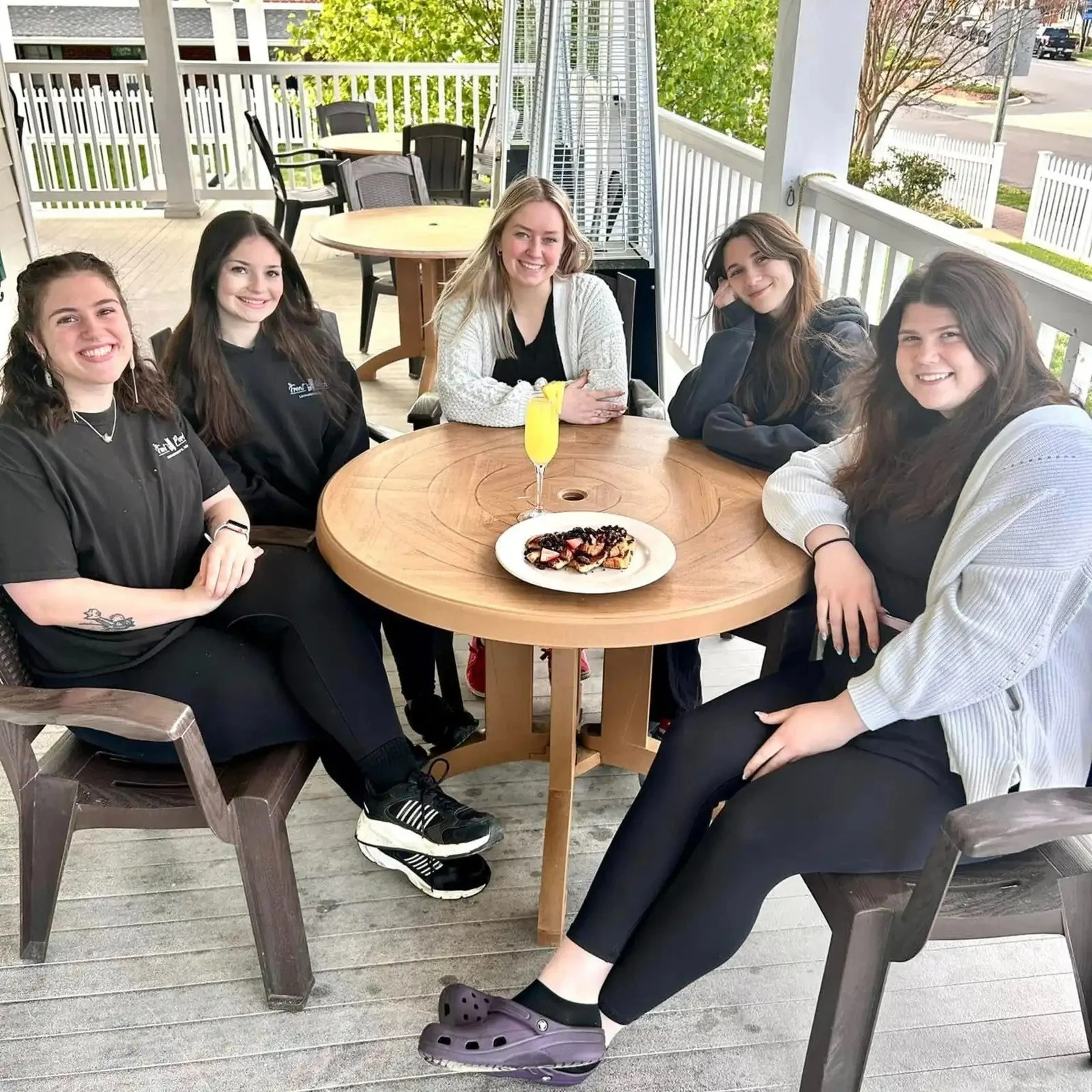 Five young women sit around a picnic table outdoors enjoying dessert and drinks on a covered porch, smiling at the camera.