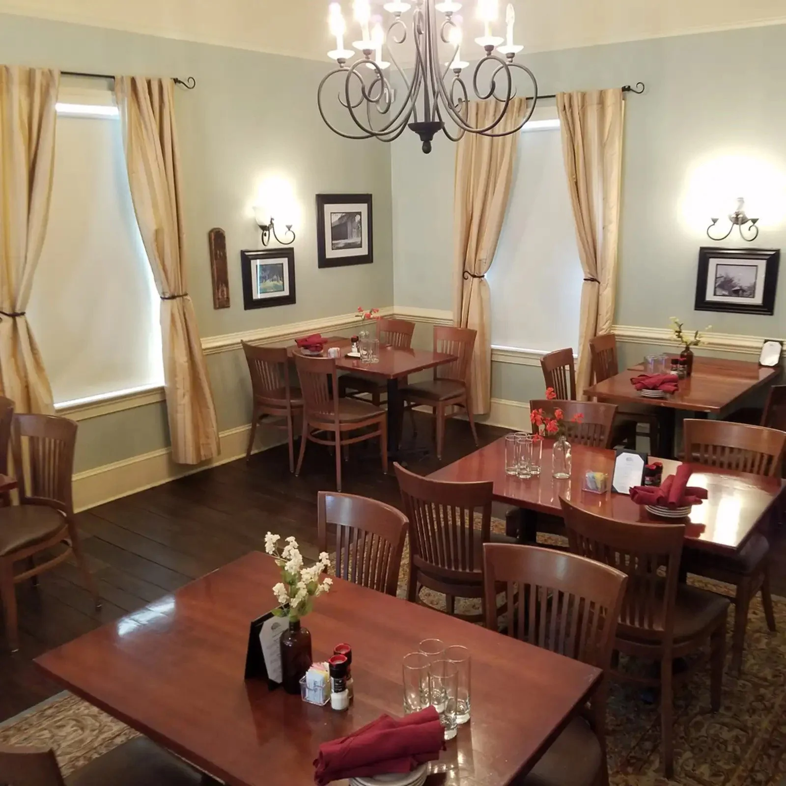 A cozy restaurant dining area with wooden tables and chairs, decorated with red napkins, flowers in vases, framed black and white artwork on mint green walls, and beige curtains hanging by windows. A chandelier hangs from the ceiling, and wall sconces provide warm lighting.