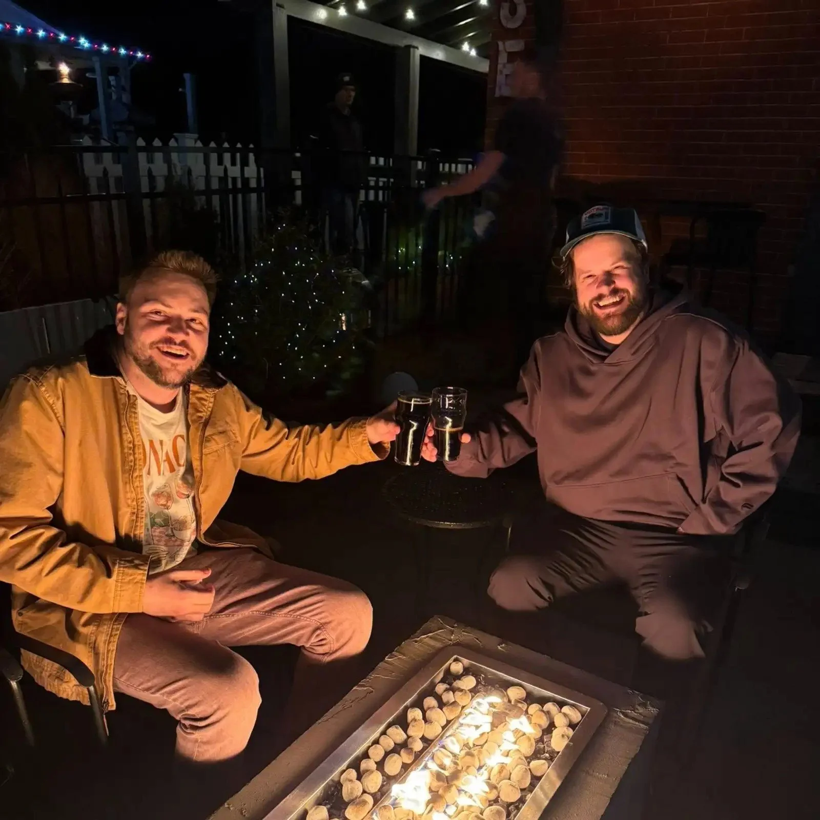 Two men sitting around a firepit, smiling and toasting drinks at night. There are decorations and other people in the background.