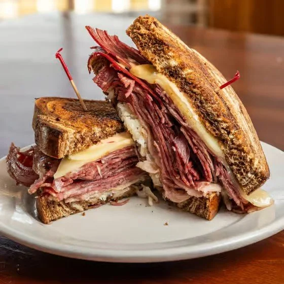 A classic Reuben sandwich cut diagonally, showing corned beef, Swiss cheese, sauerkraut, and Russian dressing on toasted rye bread, served on a white plate.