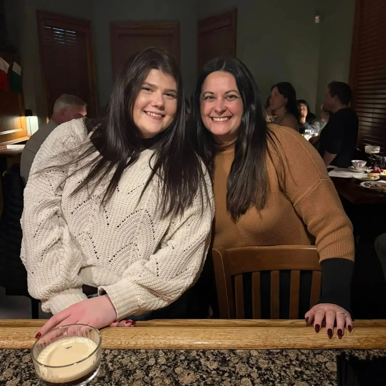 Two women sitting side by side at a restaurant table, smiling at the camera. The woman on the left has long dark hair and is wearing a cream-colored sweater, while the woman on the right has shoulder-length dark hair and is wearing a brown top. A glass candle holder with a lit candle is on the table in front of them. In the background, other diners eat and socialize in a warmly lit room.