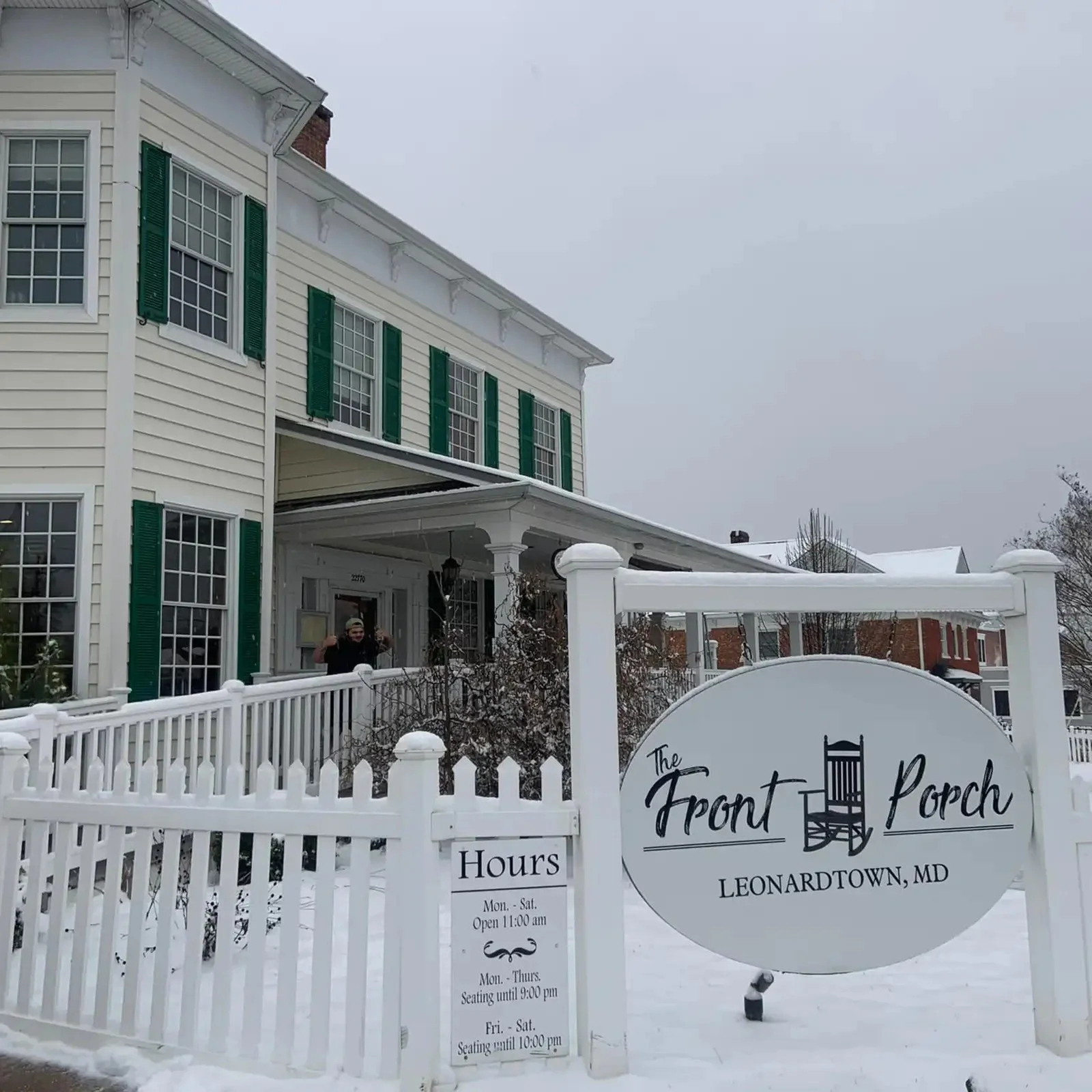 White picket fence and sign for The Front Porch restaurant in Leonardtown, Maryland, with snowy ground and part of a white house with green shutters in the background.