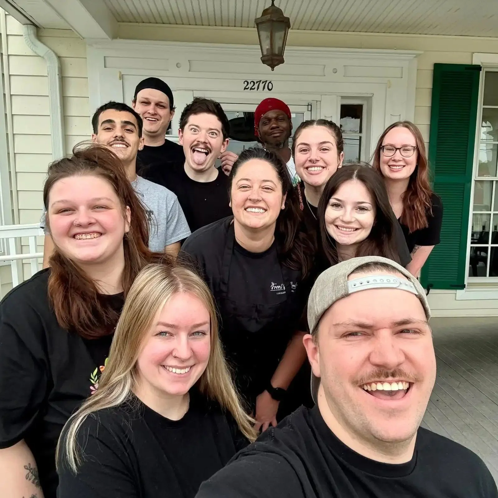Group of diverse young smiling people posing for a selfie outside a house with beige siding, green shutters, and a porch light.