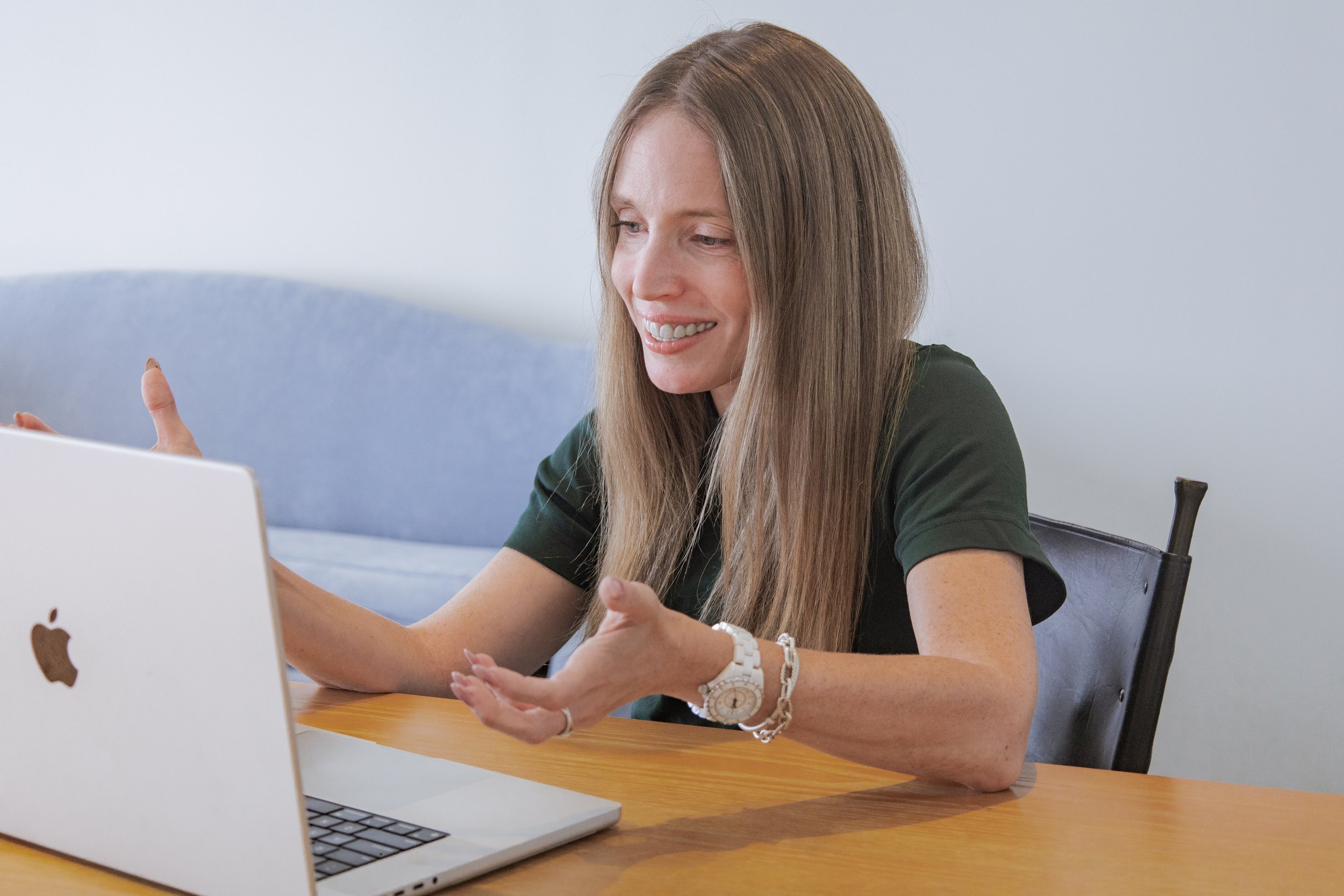 A woman with long brown hair smiling and gesturing while sitting at a wooden table with an open laptop in front of her. She is wearing a green shirt, a white watch, and bracelets.