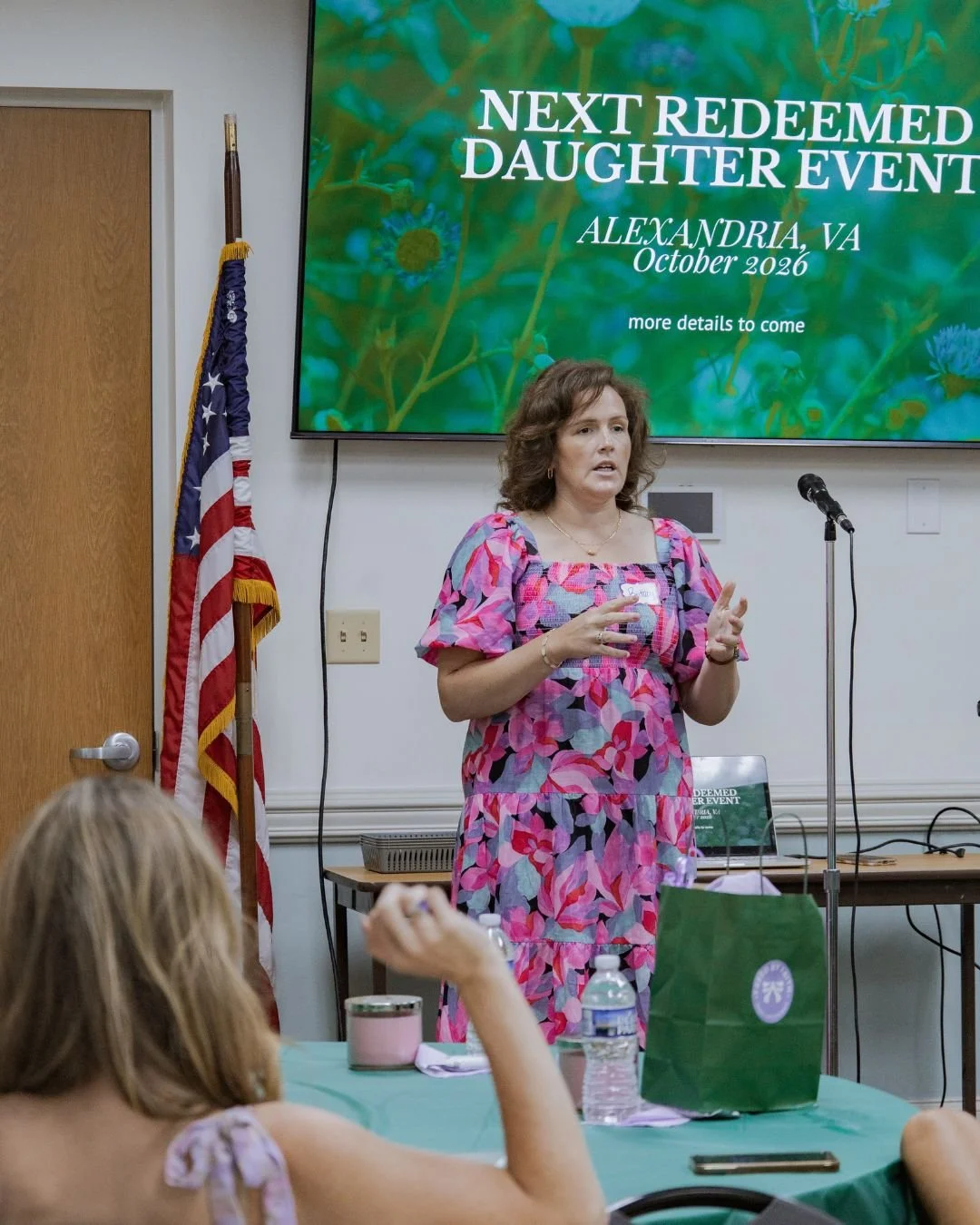A woman with curly brown hair, wearing a pink floral dress, is giving a presentation at a conference. Behind her, a large screen displays 'Next Redeemed Daughter Event, Alexandria, VA, October 2026, more details to come.' An American flag is to her left, and there is a microphone stand in front of her. The audience includes a person with long blonde hair seated at a table with a green tablecloth, a water bottle, and a pink mug.