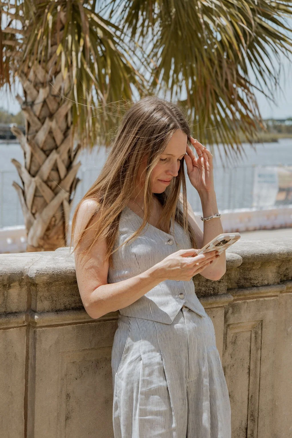 A woman with long light brown hair looking at her phone outdoors near water, with a palm tree in the background.