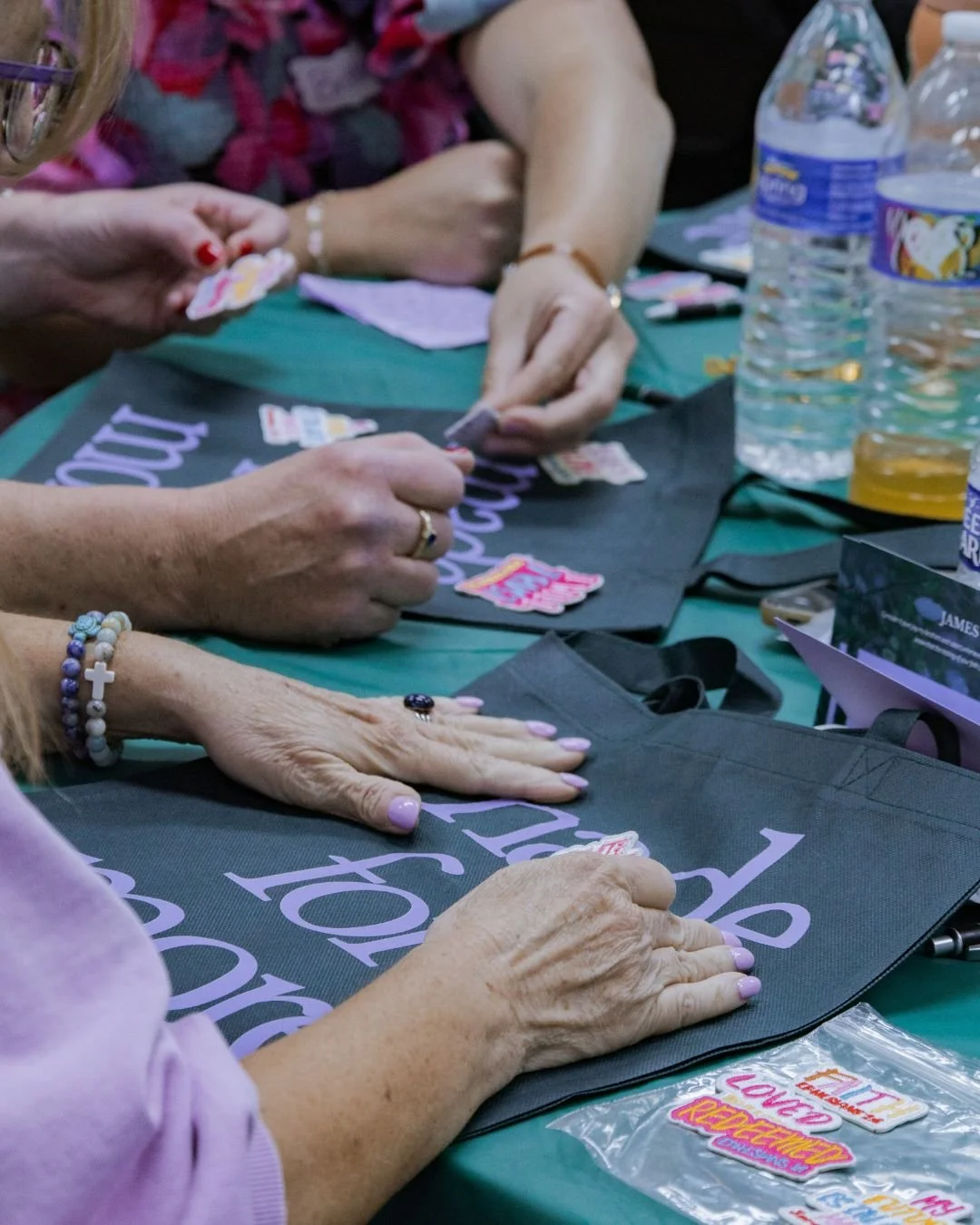 People signing stickers on black tote bags at a table with water bottles and crafting materials.