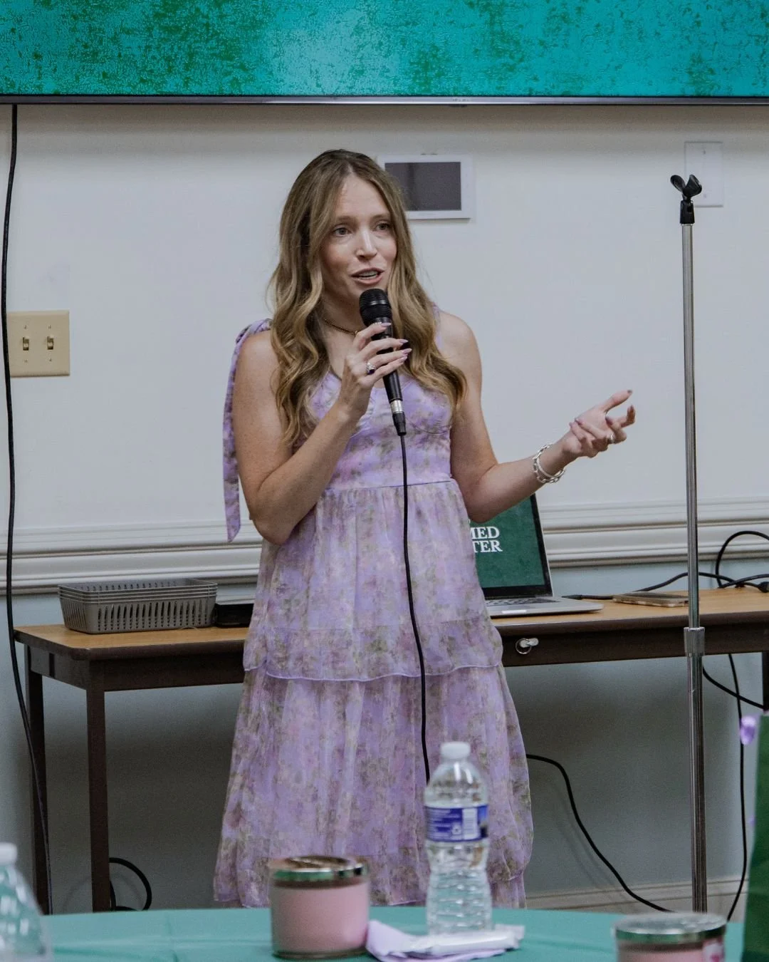 A woman with long, wavy hair wearing a light purple, patterned dress is speaking into a microphone during a presentation or speech in a room with a green and white background.