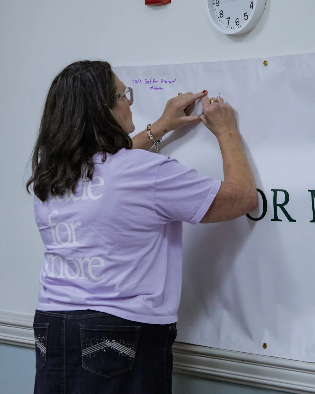 Woman writing on a large white paper on a wall with a marker, wearing a pink T-shirt that says 'guide for more'. A clock is visible on the wall above her.