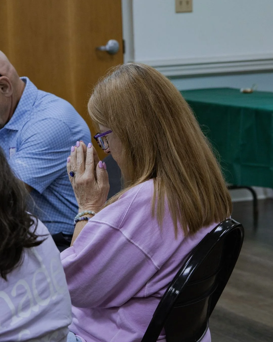 A woman with red hair wearing glasses, a pink sweatshirt, and jewelry has her hand raised with her fingers pressed together, holding her hand near her face in a praying or thoughtful posture. She is sitting beside a man in a blue shirt, in a room with a wooden door and a green table covered with a green cloth in the background.