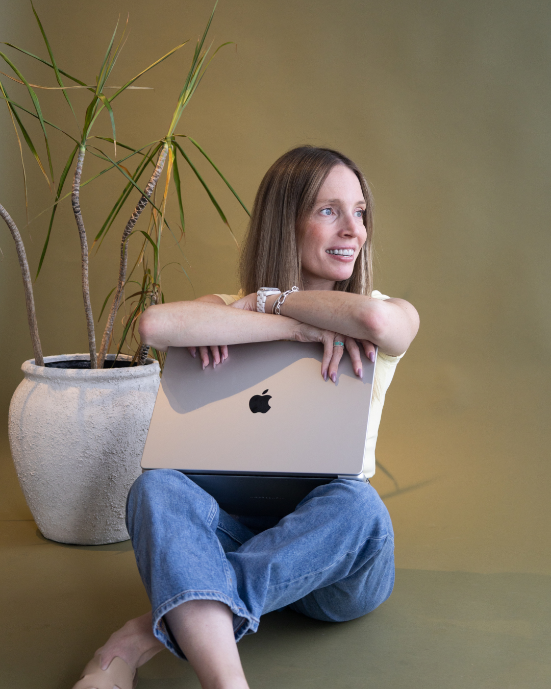 A woman with long brown hair sitting cross-legged on the floor, holding a silver MacBook laptop, smiling and looking to her right, with a large potted plant behind her against a plain beige wall.