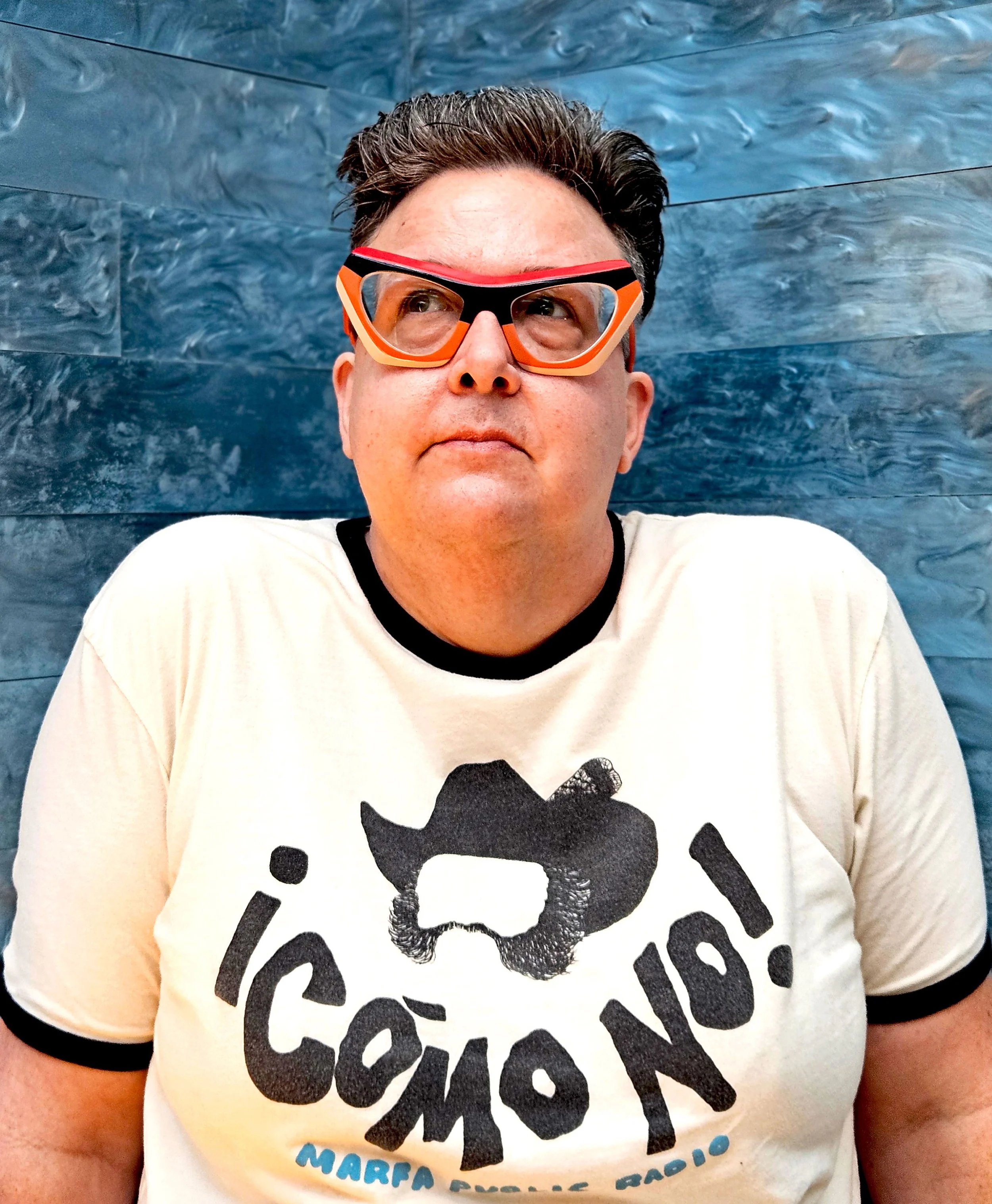 Close-up of a person's face wearing red and orange goggles, with dark hair, against a blue textured background, wearing a white T-shirt with a black skull graphic and black text.