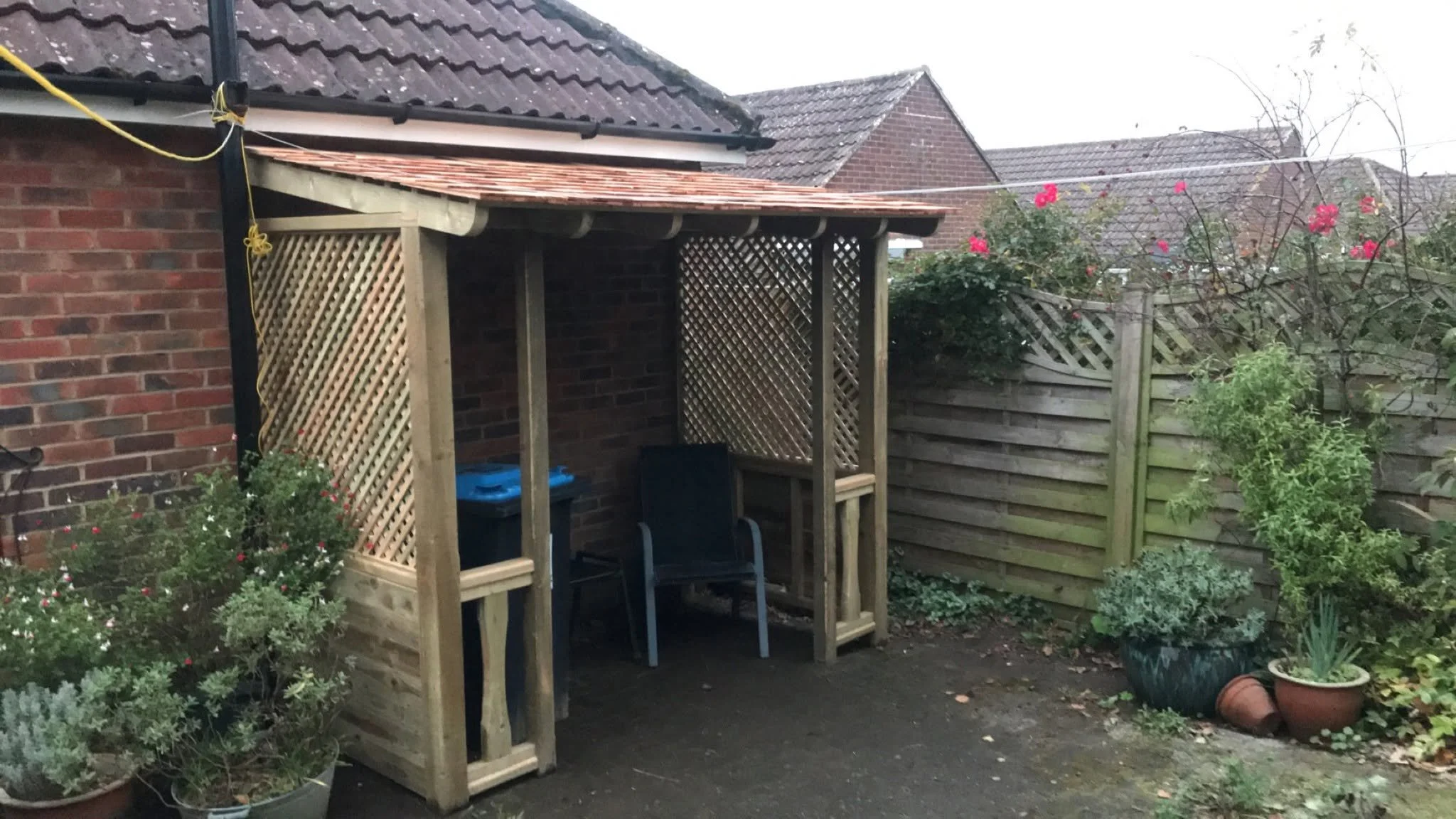 A wooden garden structure built up against the side of a garage with chairs and a table inside.
