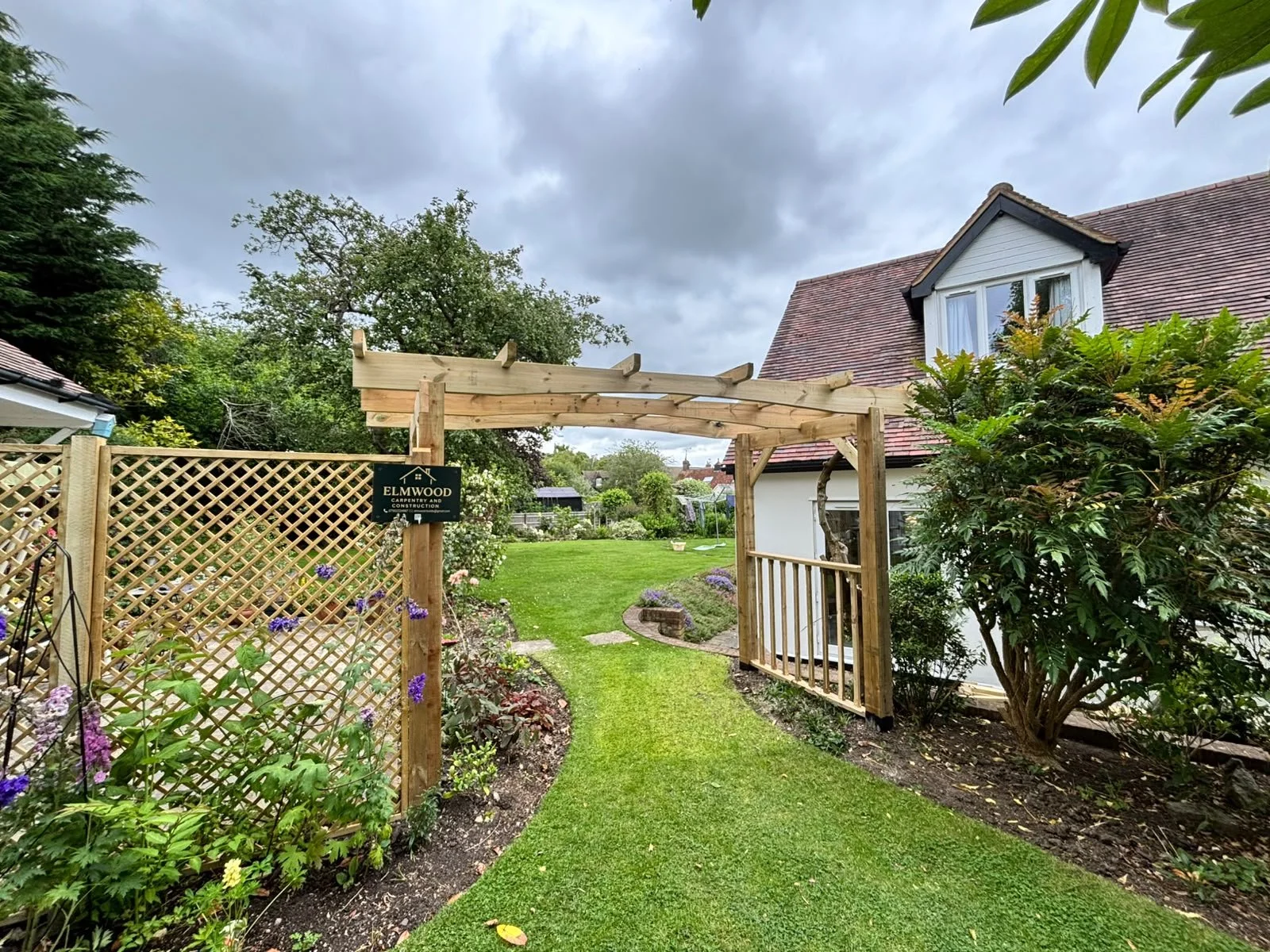 A back garden with a lawn, flower beds, and a wooden pergola, with a sign that reads 'Elmwood Carpentry and Construction.'