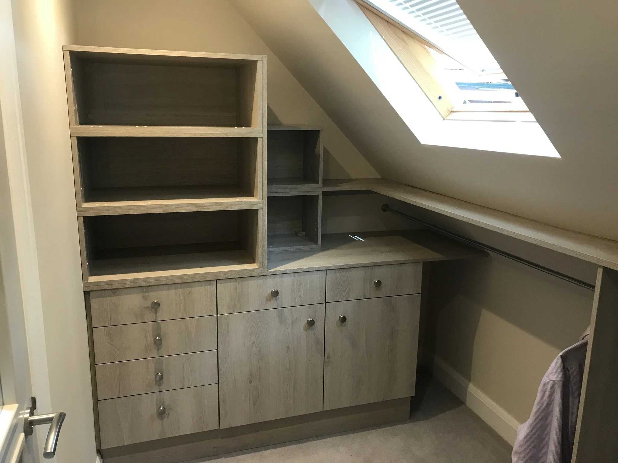 A built-in wooden cabinet with multiple open shelves and drawers, located in an attic space under a sloped ceiling with a skylight window.