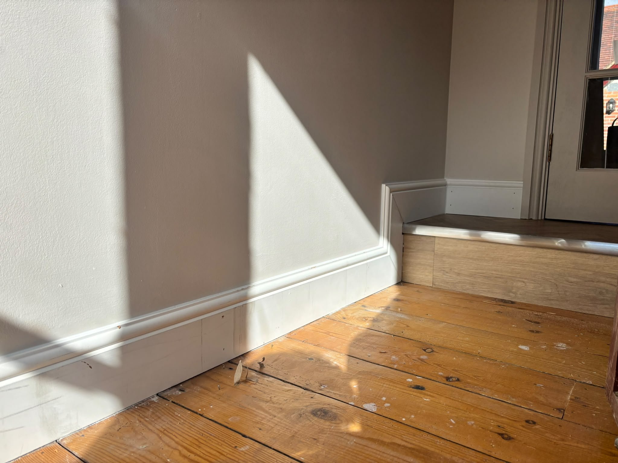 View of a corner in a house with wooden floor, beige walls, white skirting board.
