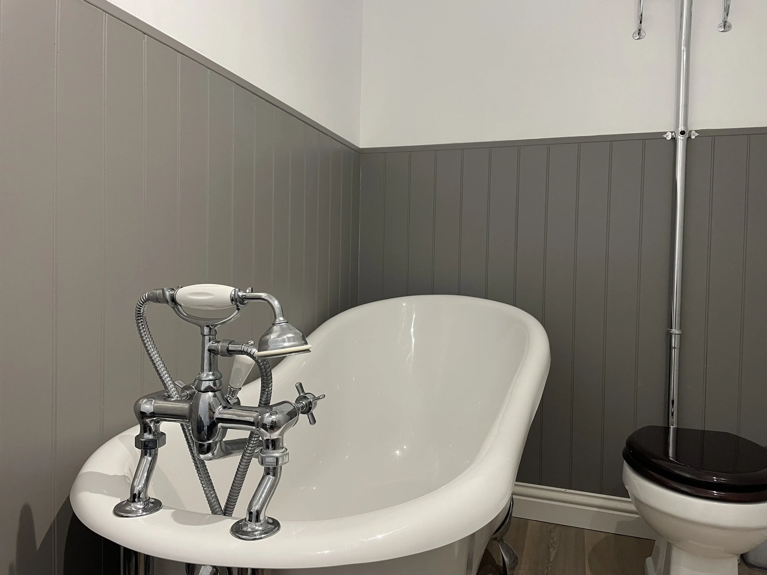 A vintage freestanding bathtub with shiny silver fixtures, a black toilet to the right, and grey wood paneling wall.
