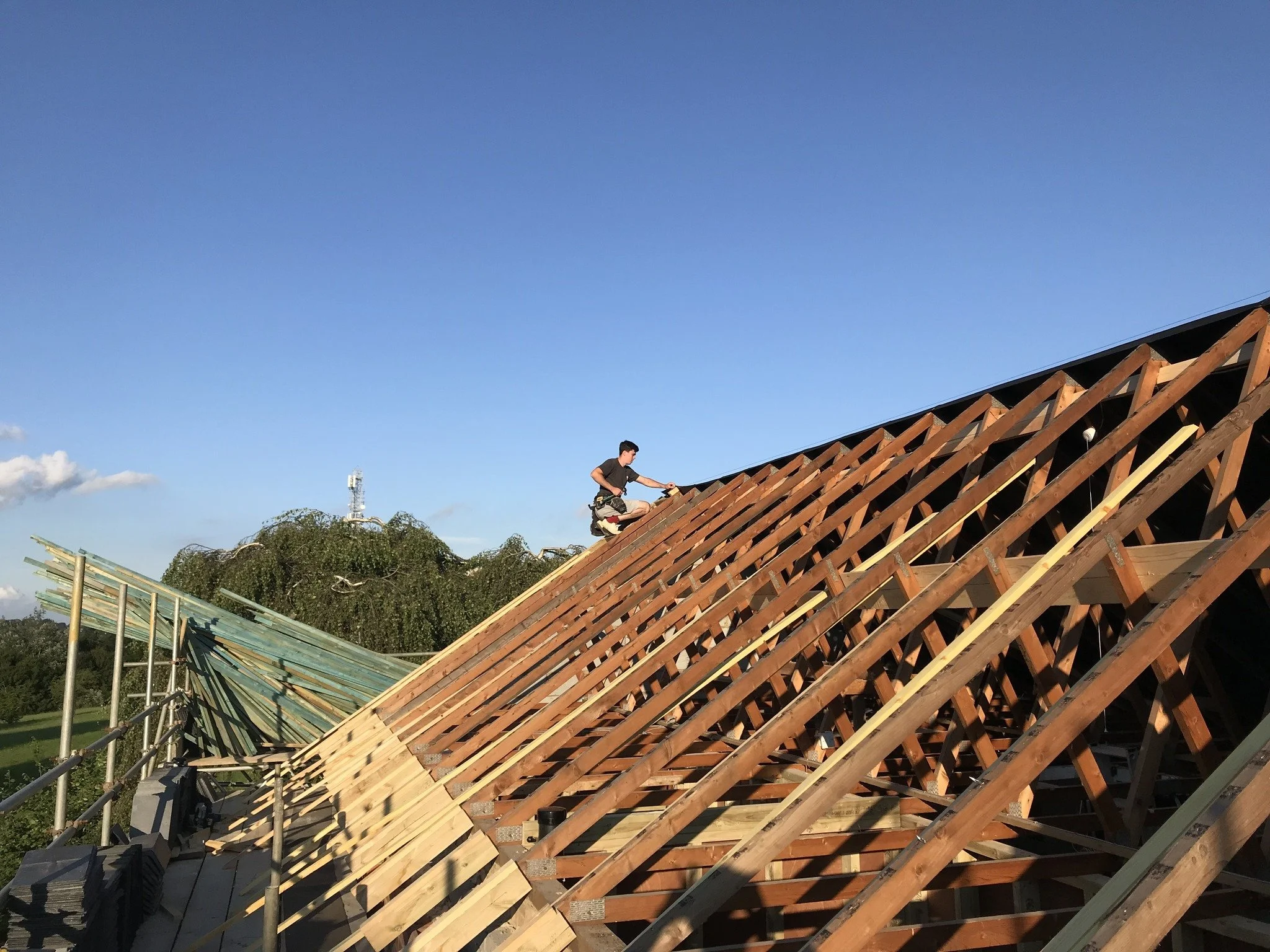 A person working on the wooden frame of a building under construction, with a clear blue sky in the background.