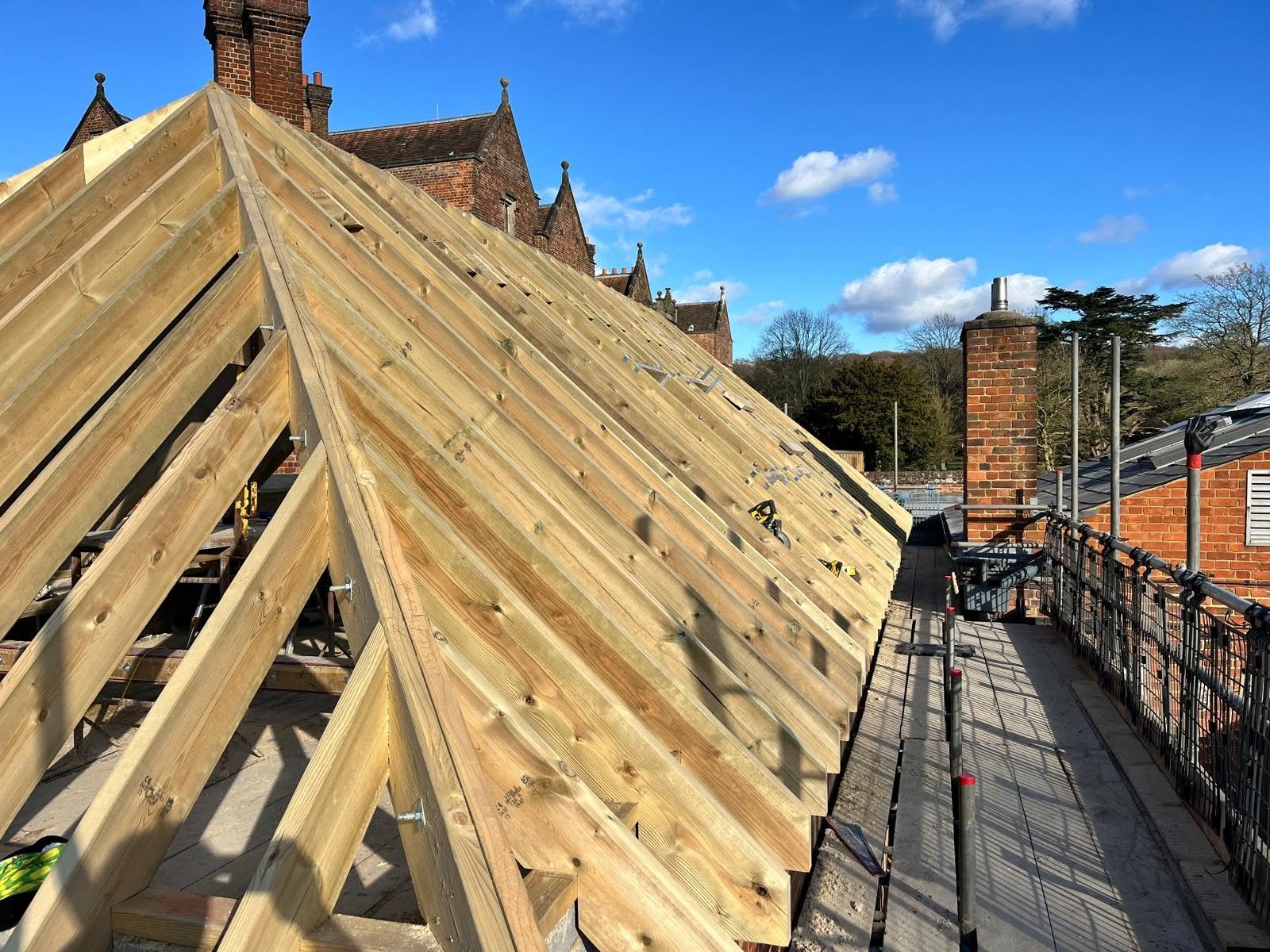 Construction site on a building rooftop with a new wooden roof frame, scaffolding, brick chimney, and a clear blue sky with a few clouds.