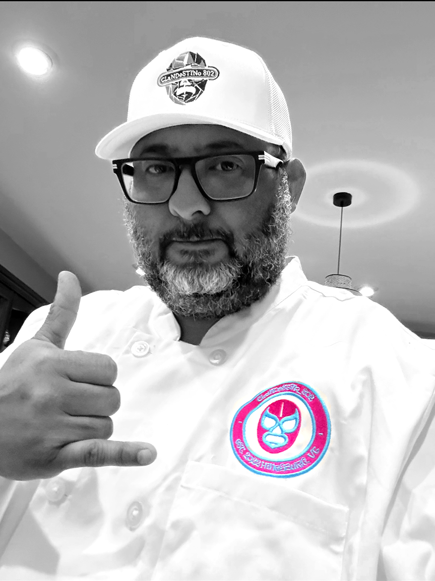 Man with glasses and a beard taking a selfie. Wearing a white chef's coat with a colorful logo on the chest, a white cap with a logo, and making a shaka sign with his hand. The background shows indoor lighting and ceiling fixtures.