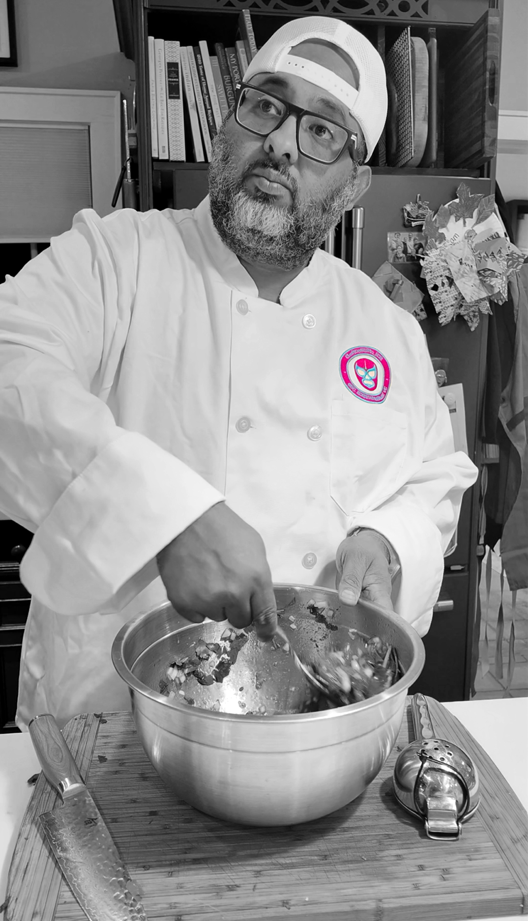 A man with glasses and a beard wearing a chef's coat and baseball cap, mixing ingredients in a large stainless steel bowl in a kitchen surrounded by books and decorations.