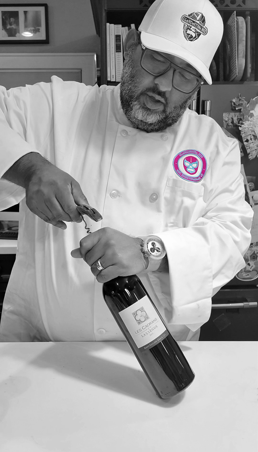A man in a chef's coat and cap opening a bottle of wine, with books and decorations in the background.