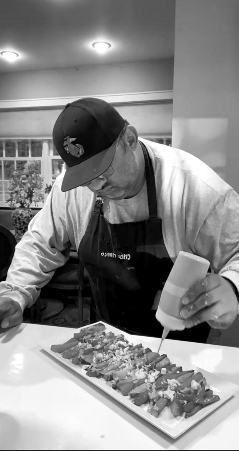 A man wearing a baseball cap, glasses, and an apron is adding sauce to a platter of food. The food appears to be sushi rolls garnished with toppings.