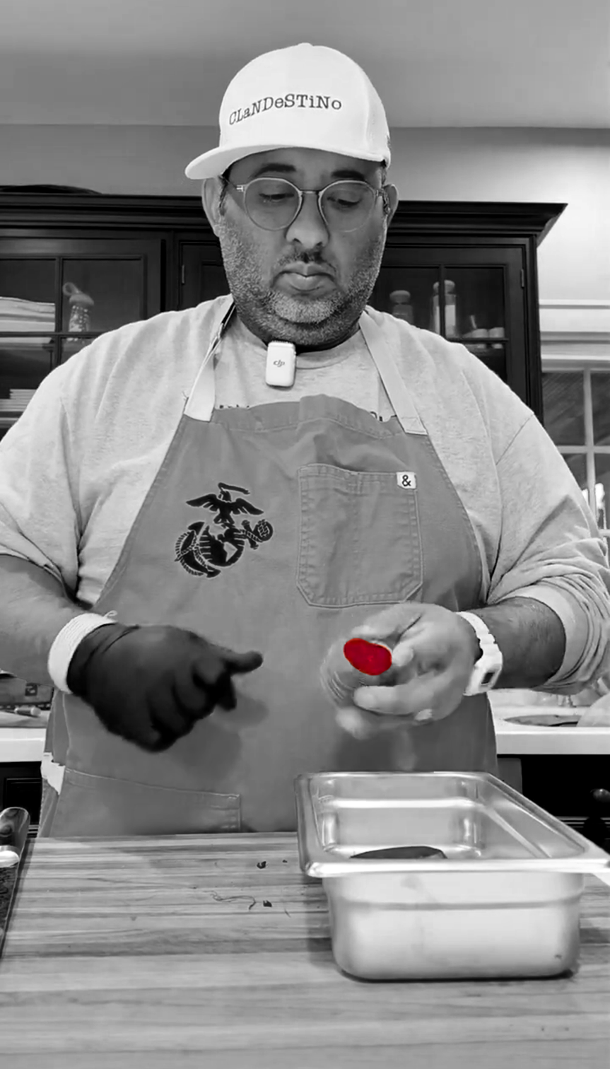 A man wearing glasses, a "Clandestino" cap, gloves, and an apron decorated with a Marine emblem is preparing food in a kitchen. He holds a red item in his hand.
