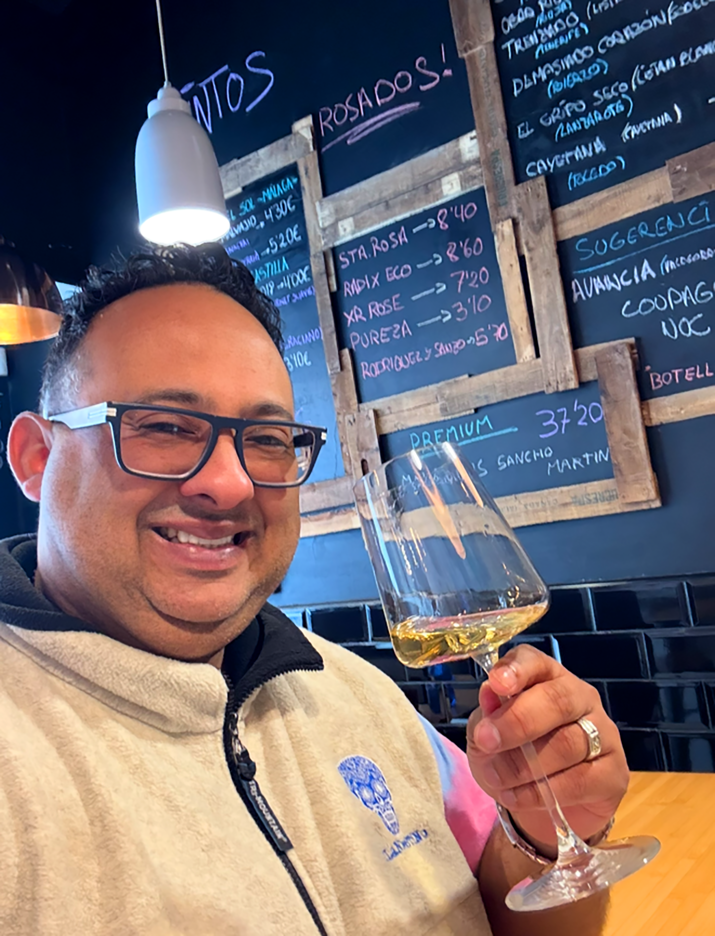 Smiling man wearing glasses and a hoodie, holding a glass of white wine in a restaurant or bar with blackboards of menu items in the background.