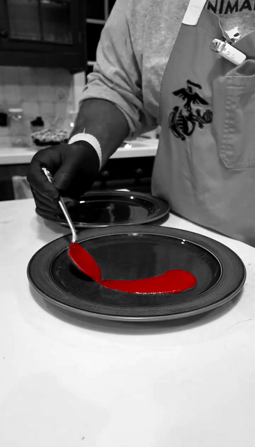 Person in a gray jacket wearing a black glove and a white wristband, filling a black plate with red sauce in a kitchen.