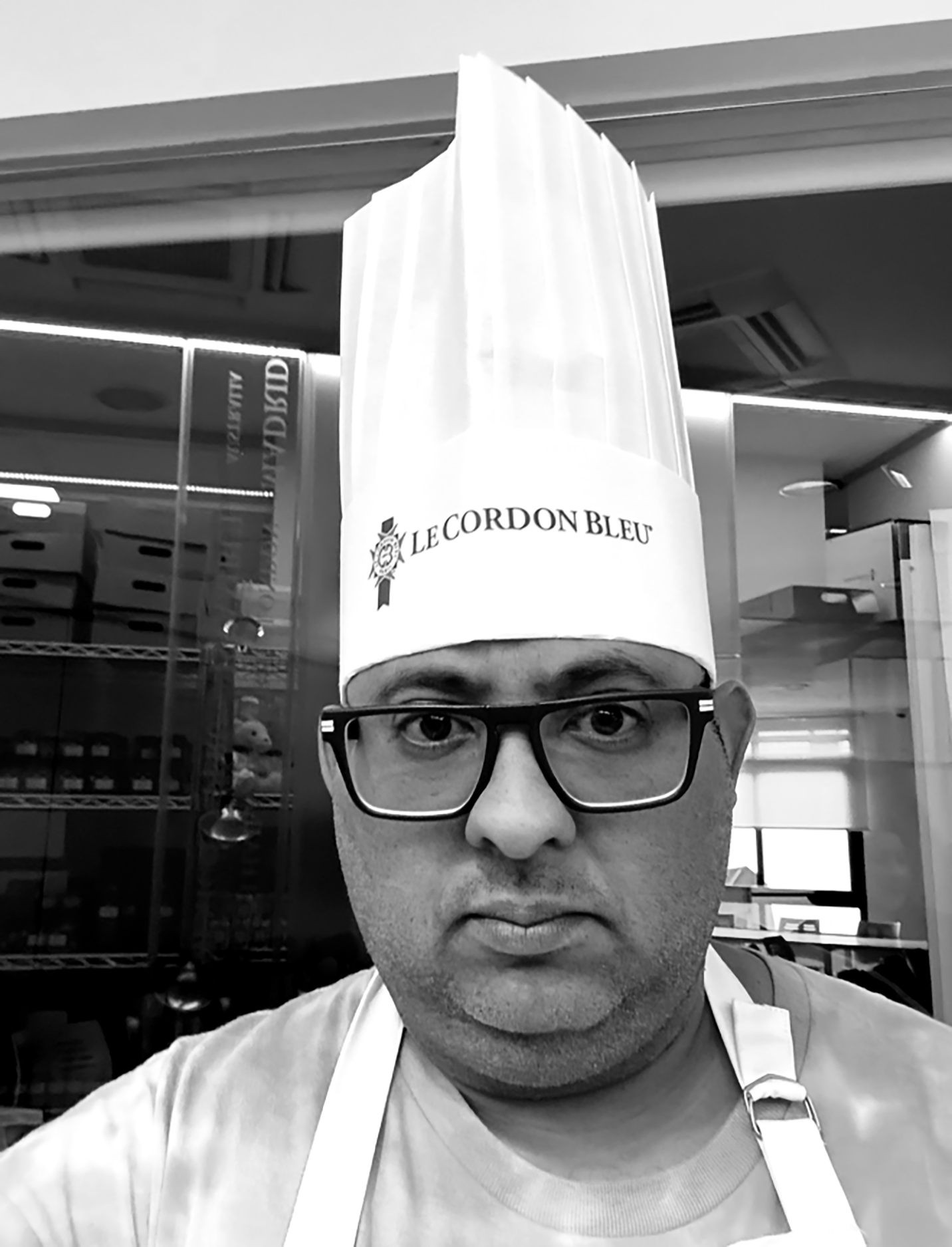 A person wearing glasses, a white apron, and a chef hat with 'Le Cordon Bleu' logo, taking a selfie in a kitchen or culinary school setting.