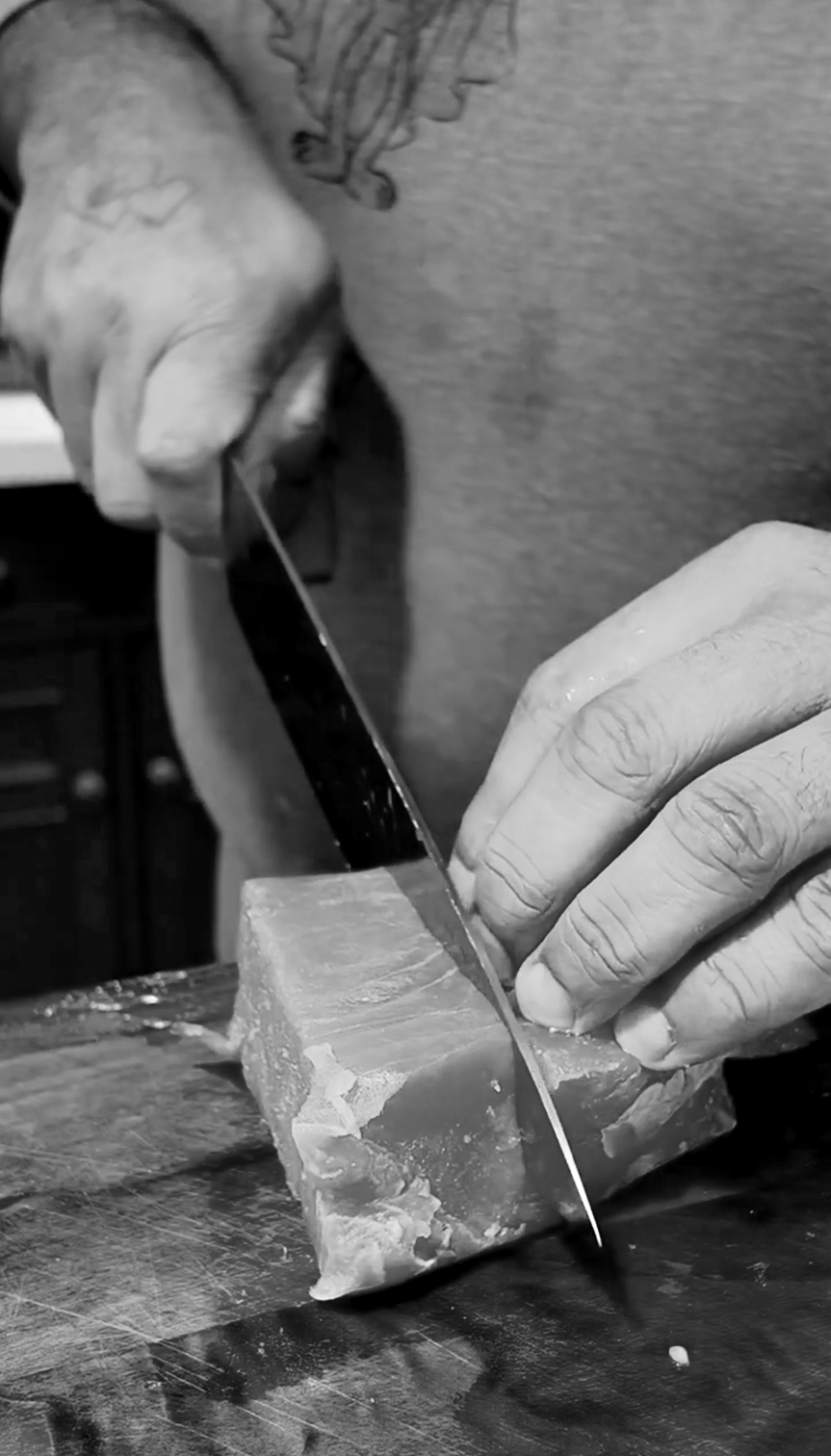 Close-up of a person's hand cutting a block of meat with a knife on a wooden cutting board.