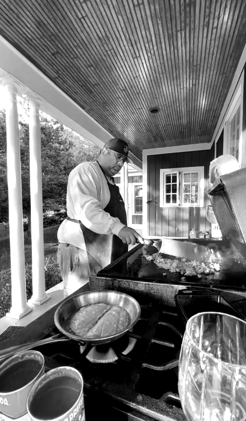 A man cooking breakfast on a grill outdoors, with a skillet and cans of food nearby.