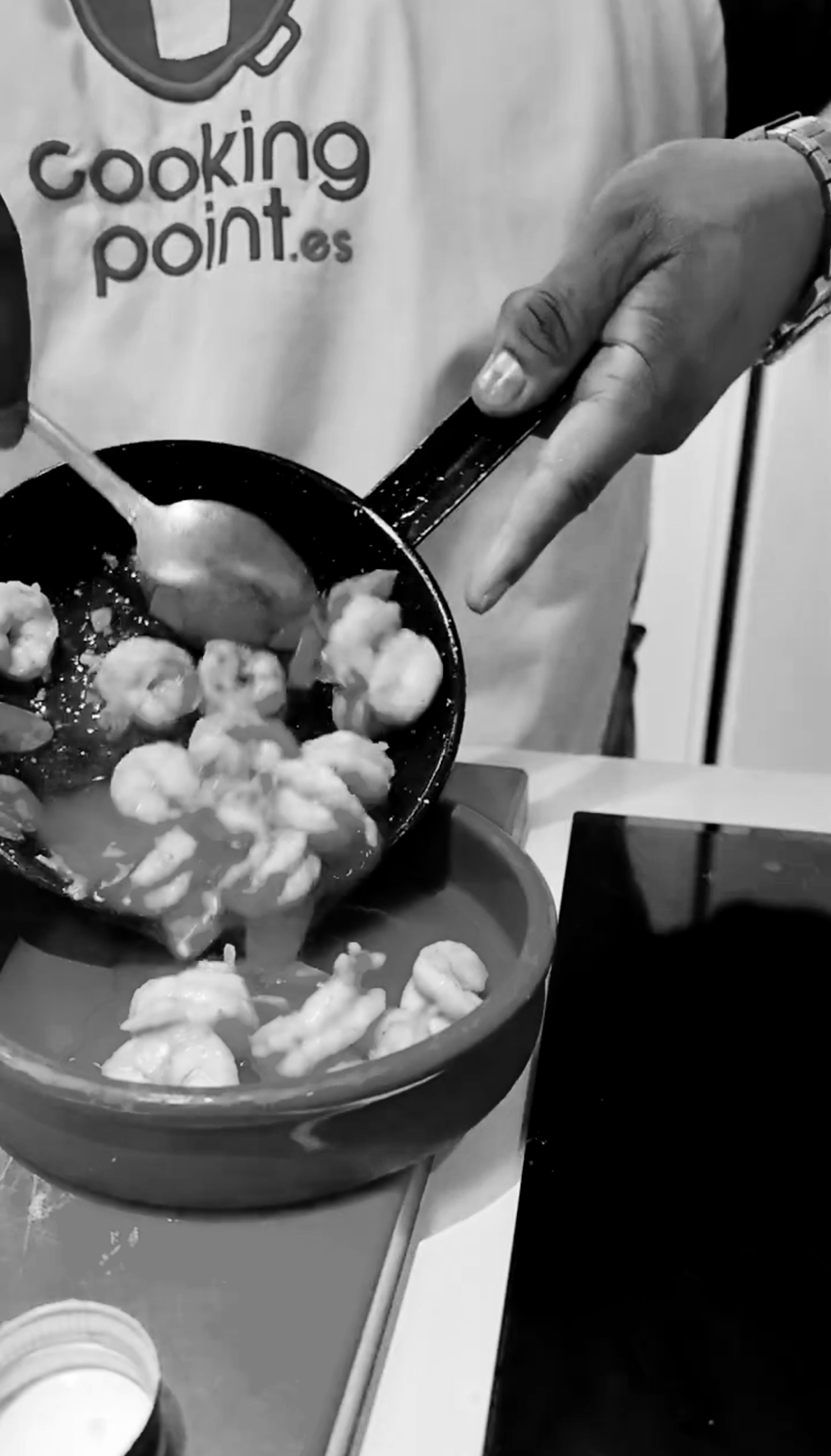 Person cooking shrimp in a frying pan over a bowl, wearing an apron with 'cookingpoint.es' written on it.