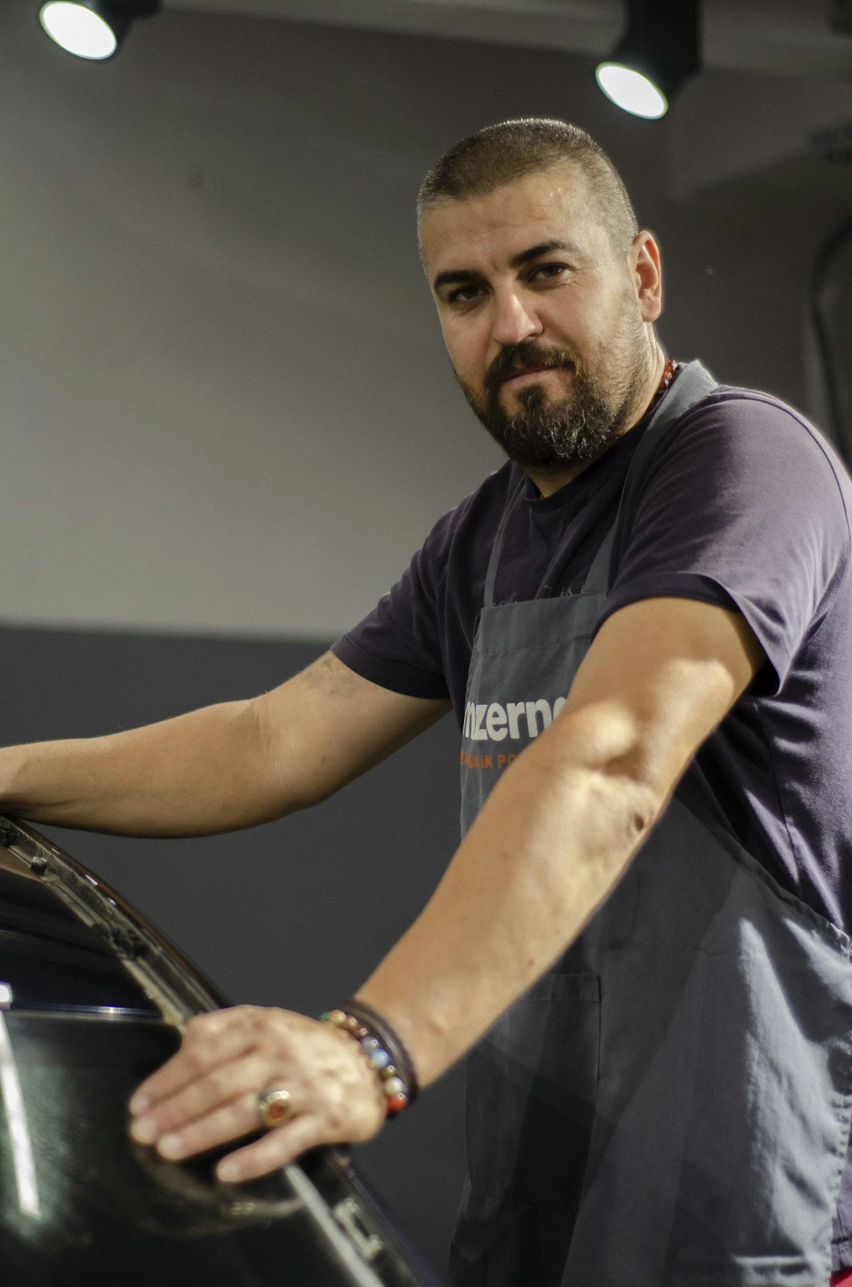 A man with a beard and short hair, wearing a dark shirt and a gray apron, is working on a cooking or grilling station. He has tattoos on his forearm and is focused on his task, with a dark background and ceiling lights above.