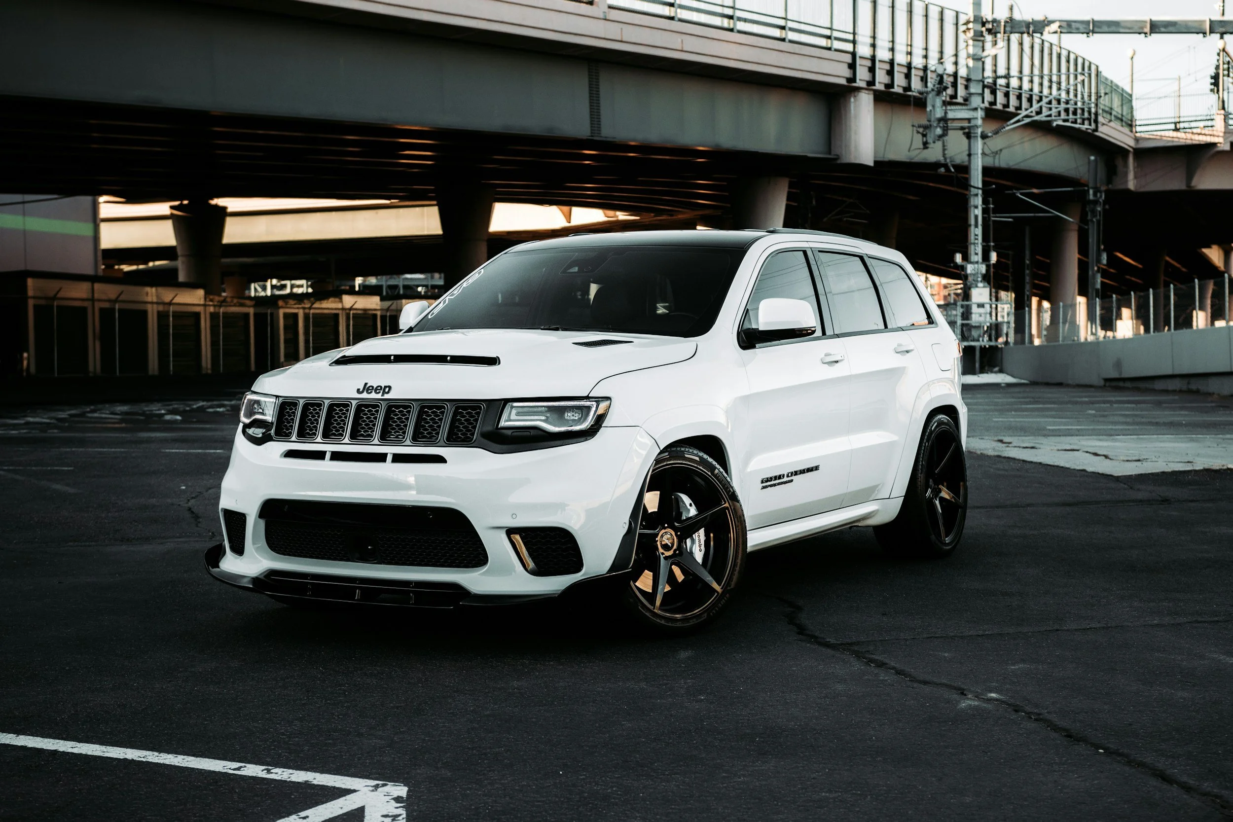 A white Jeep SUV parked in an urban parking lot under a multi-level parking structure with dark tinted windows and black wheels.