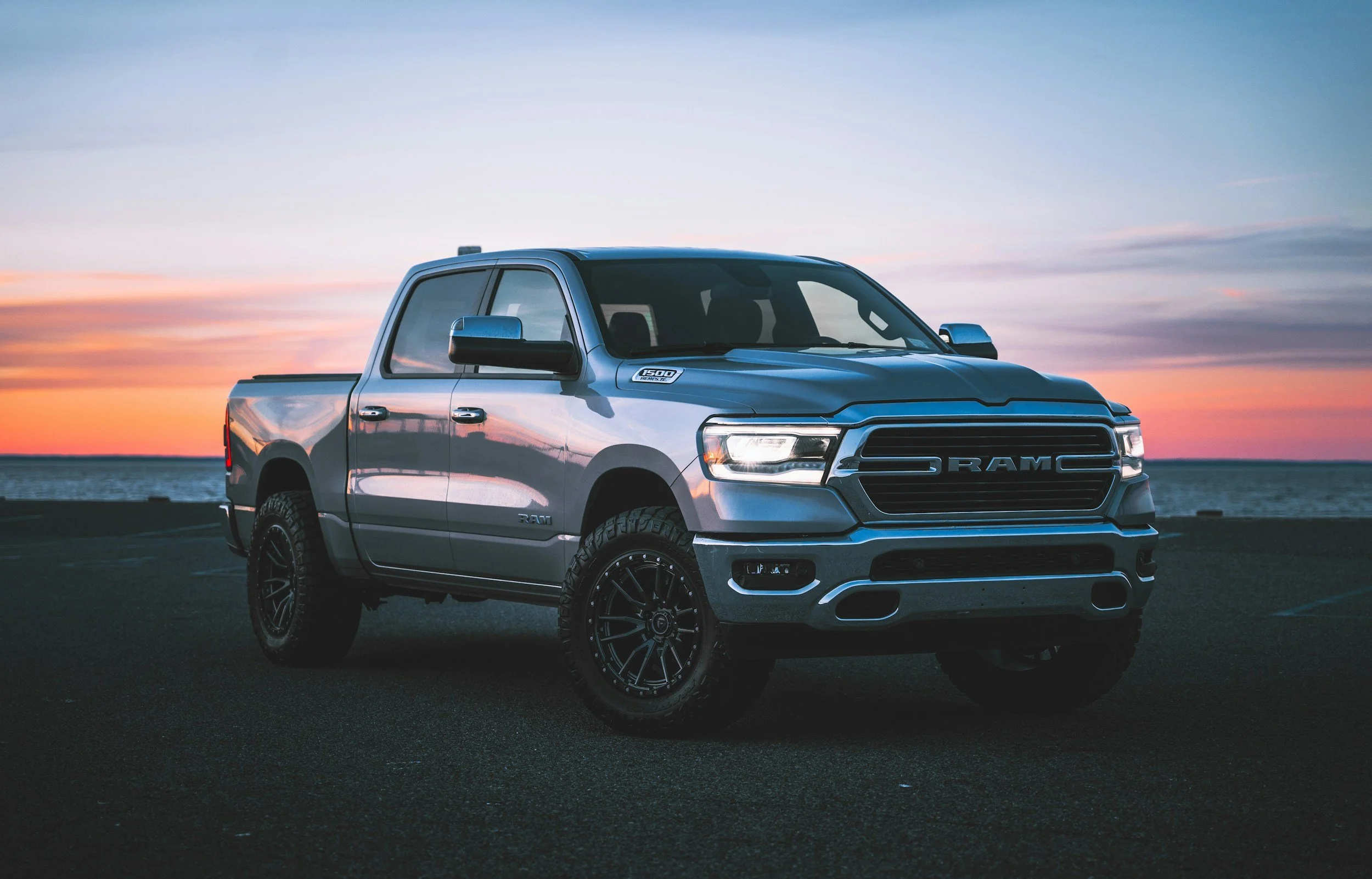 A silver Ram 1500 pickup truck parked outdoors near a body of water during sunset, with a colorful sky and calm water in the background.