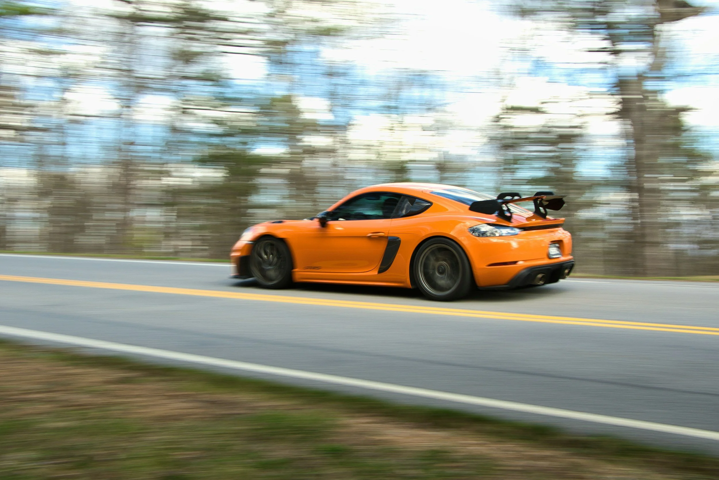 An orange sports car with black wheels and a rear spoiler driving fast on a highway, with trees blurred in the background due to motion.