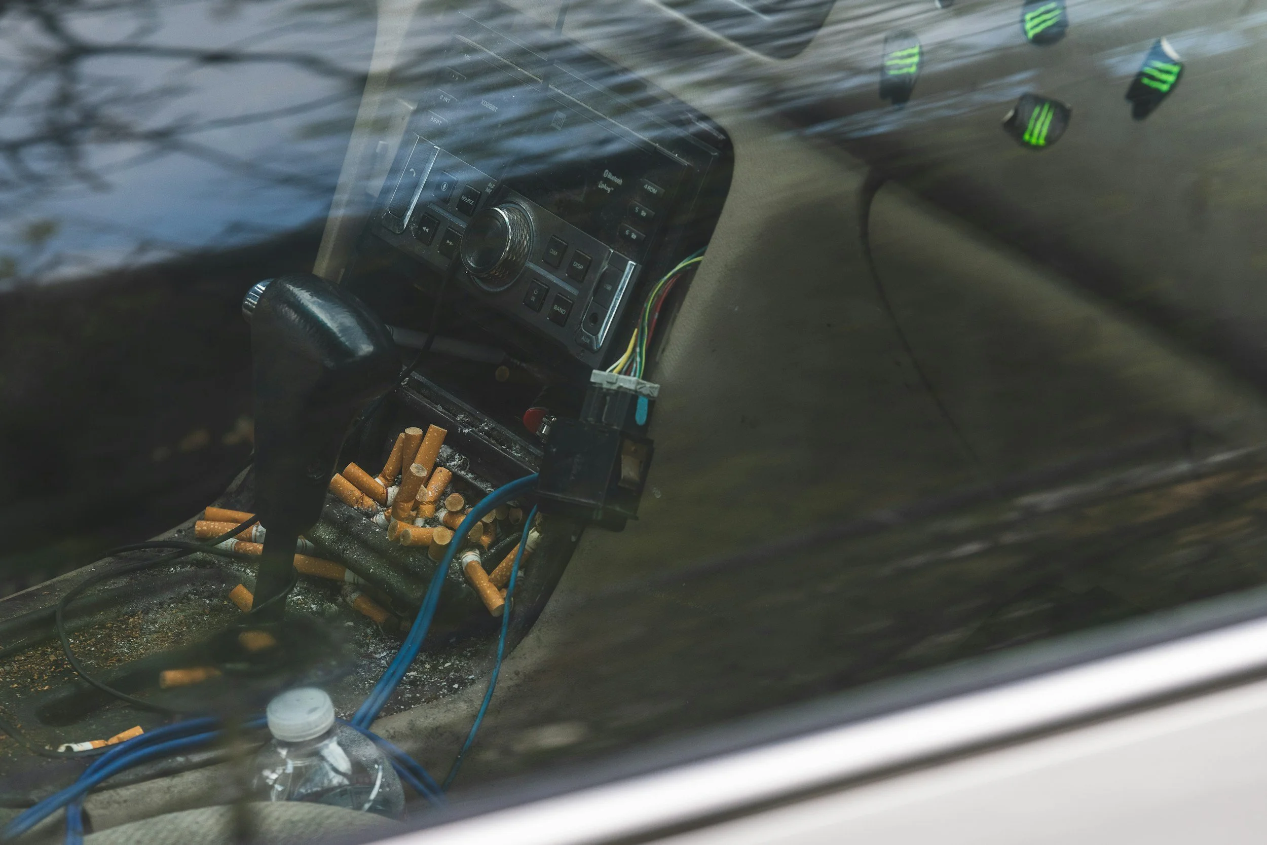 The image shows the view through a car windshield, featuring a car stereo system, several cigarette butts, a water bottle, and various wires, with some reflections on the glass.