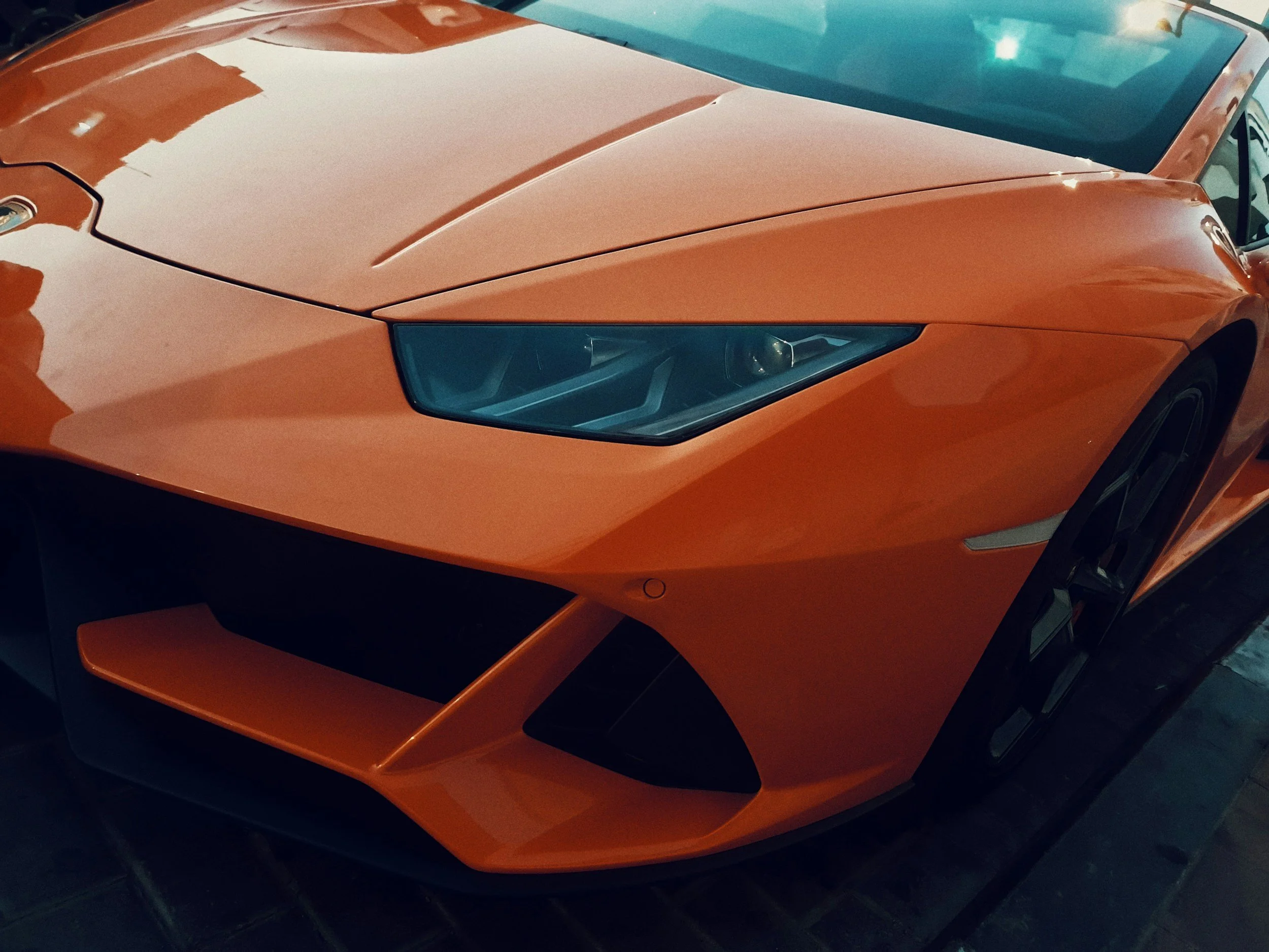 Close-up of an orange sports car with sharp, angular design and sleek headlight, parked indoors on a tiled floor.