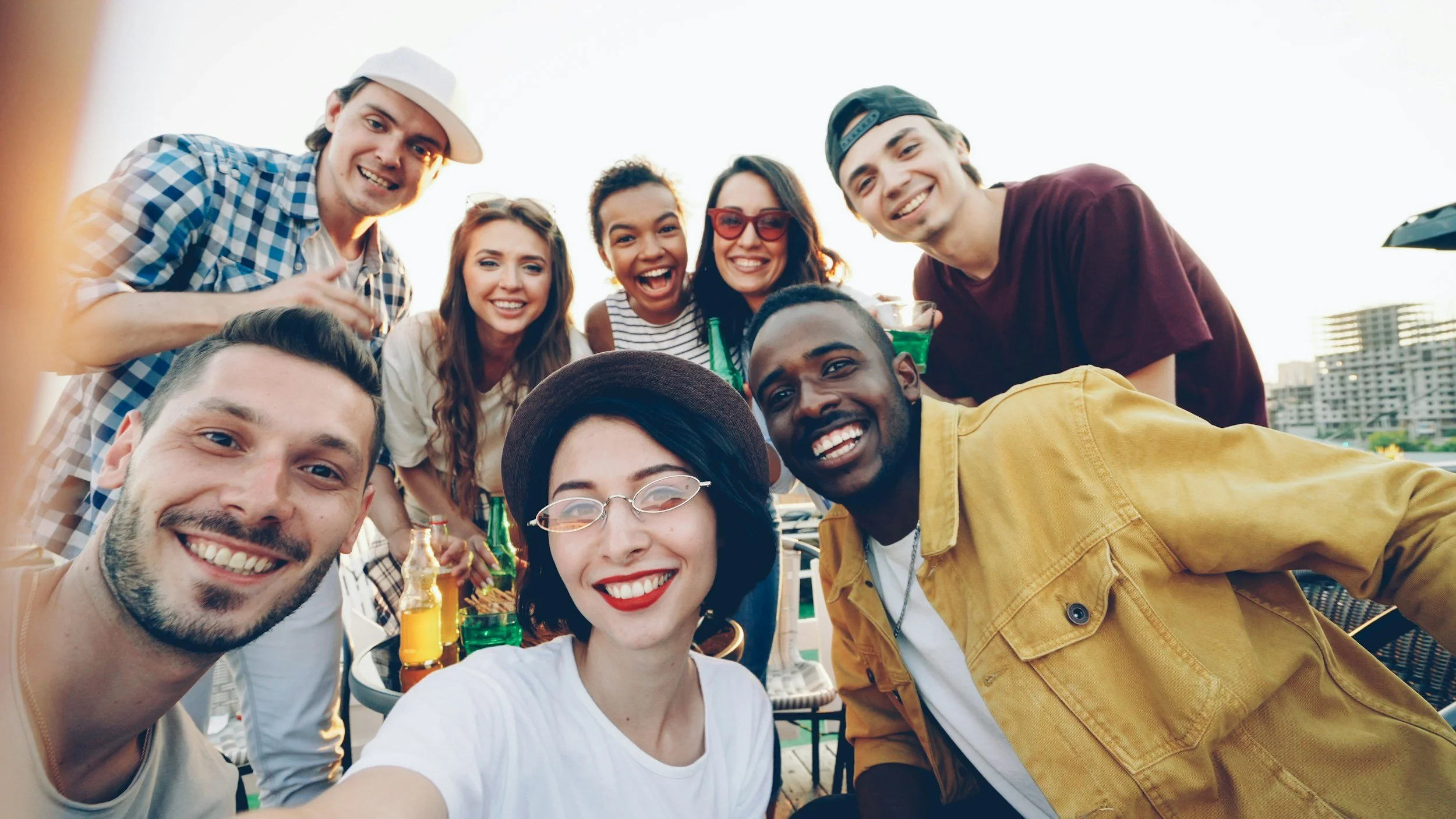 Group of friends smiling and taking a selfie at an outdoor gathering with drinks, city buildings in the background.