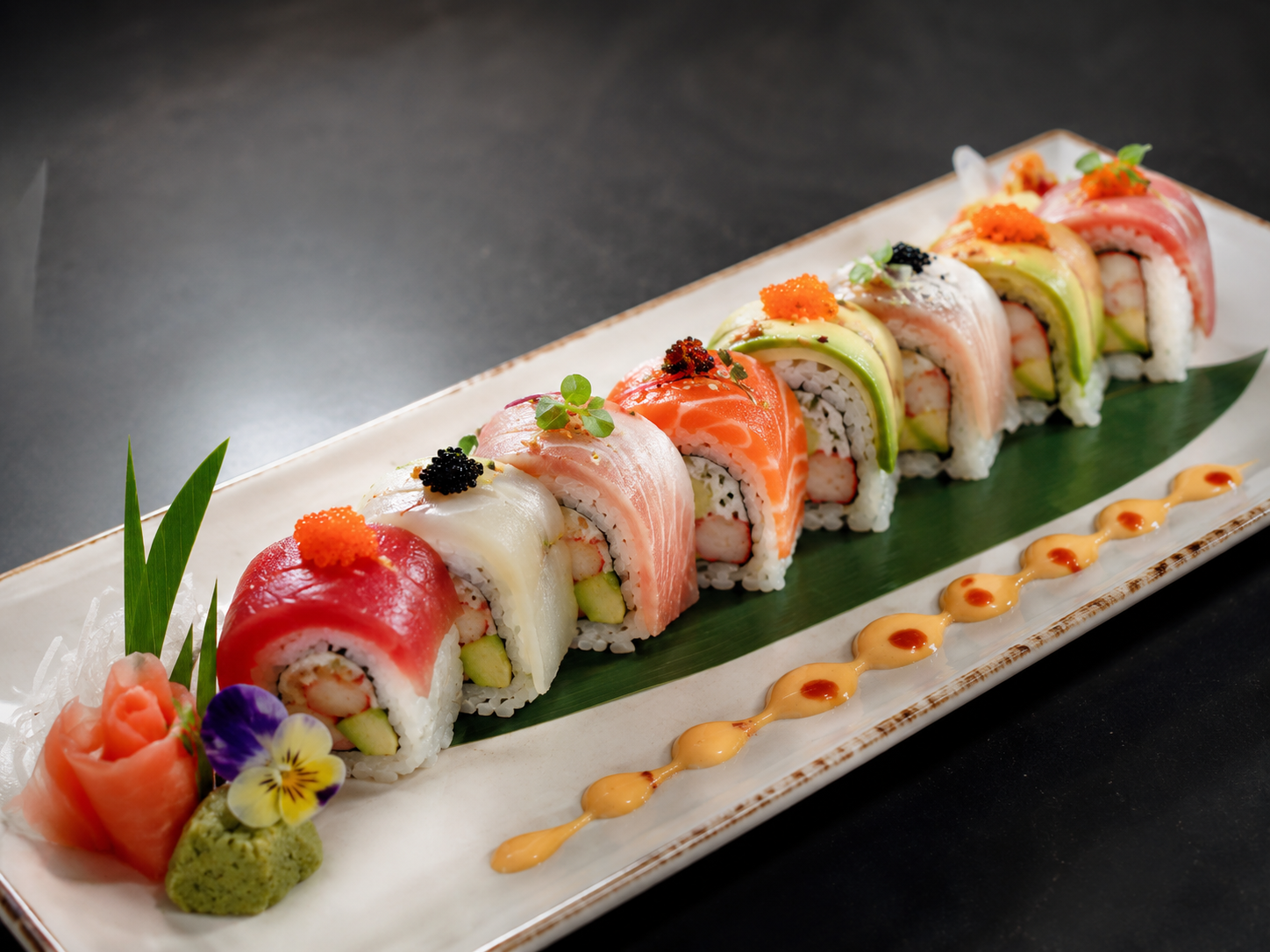 Colorful assorted sushi rolls on a rectangular white plate decorated with a green leaf, wasabi, pickled ginger, and edible flowers, with a sauce drizzle.